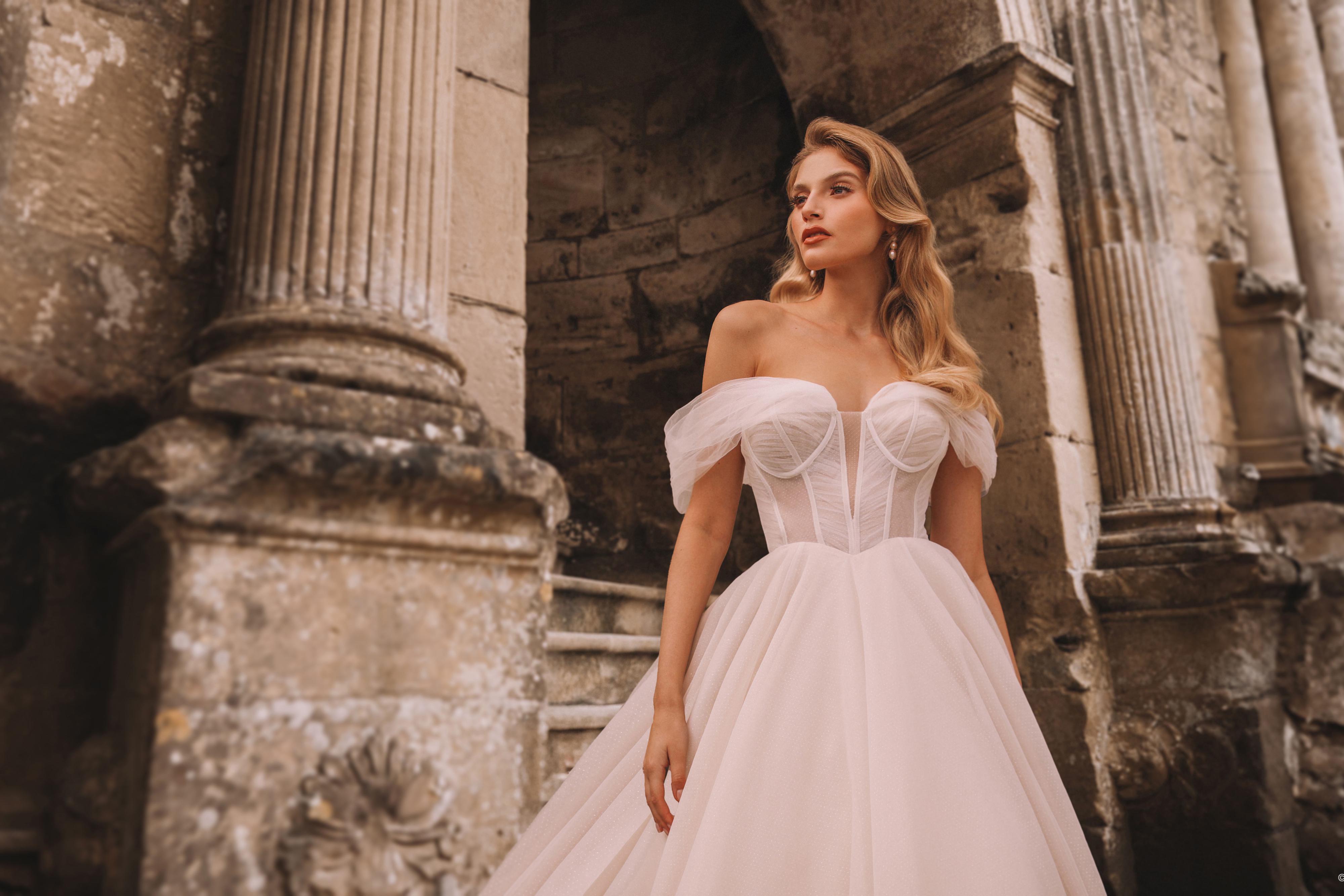 The image shows a woman wearing a stunning and elegant white wedding gown. This shows a close up of the bodice of the dress. The dress features an off-the-shoulder neckline with delicate, slightly ruffled sleeves, creating a romantic and graceful look. The bodice is fitted, enhancing the waist, and the skirt flows into a full, voluminous design that extends into a long, dramatic train behind her. The fabric appears to be light and airy, giving the gown a soft and ethereal quality. The woman's hair is styled in loose waves, complementing the overall romantic and timeless appearance of the dress.