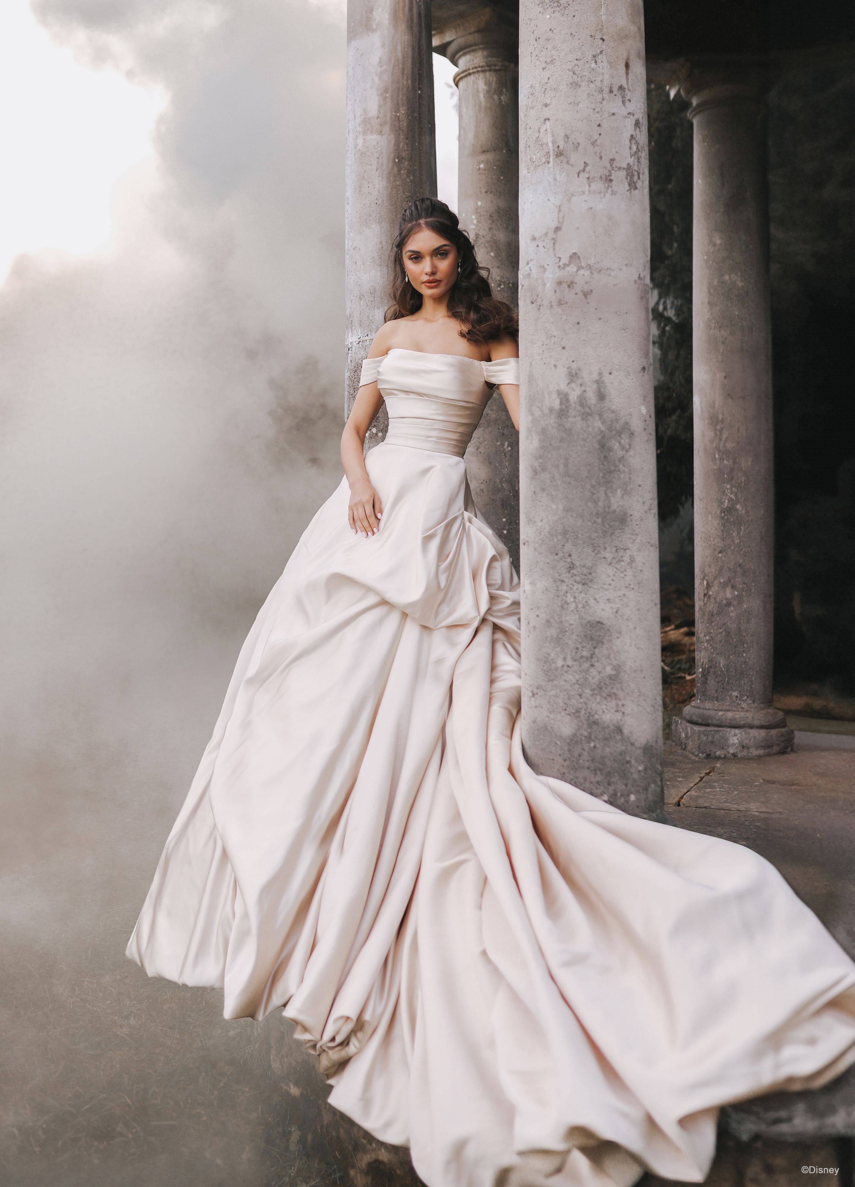 The image shows a woman wearing a luxurious and dramatic white wedding gown. The dress features an off-the-shoulder neckline and a fitted bodice that accentuates the waist. The skirt is exceptionally full and voluminous, with elaborate draping and folds that create a grand and opulent silhouette. The fabric appears to be heavy and rich, contributing to the gown's regal and elegant appearance. The long train flows behind her, adding to the overall majestic and sophisticated look of the dress. The woman's hair is styled in loose waves, enhancing the romantic and timeless quality of the ensemble.