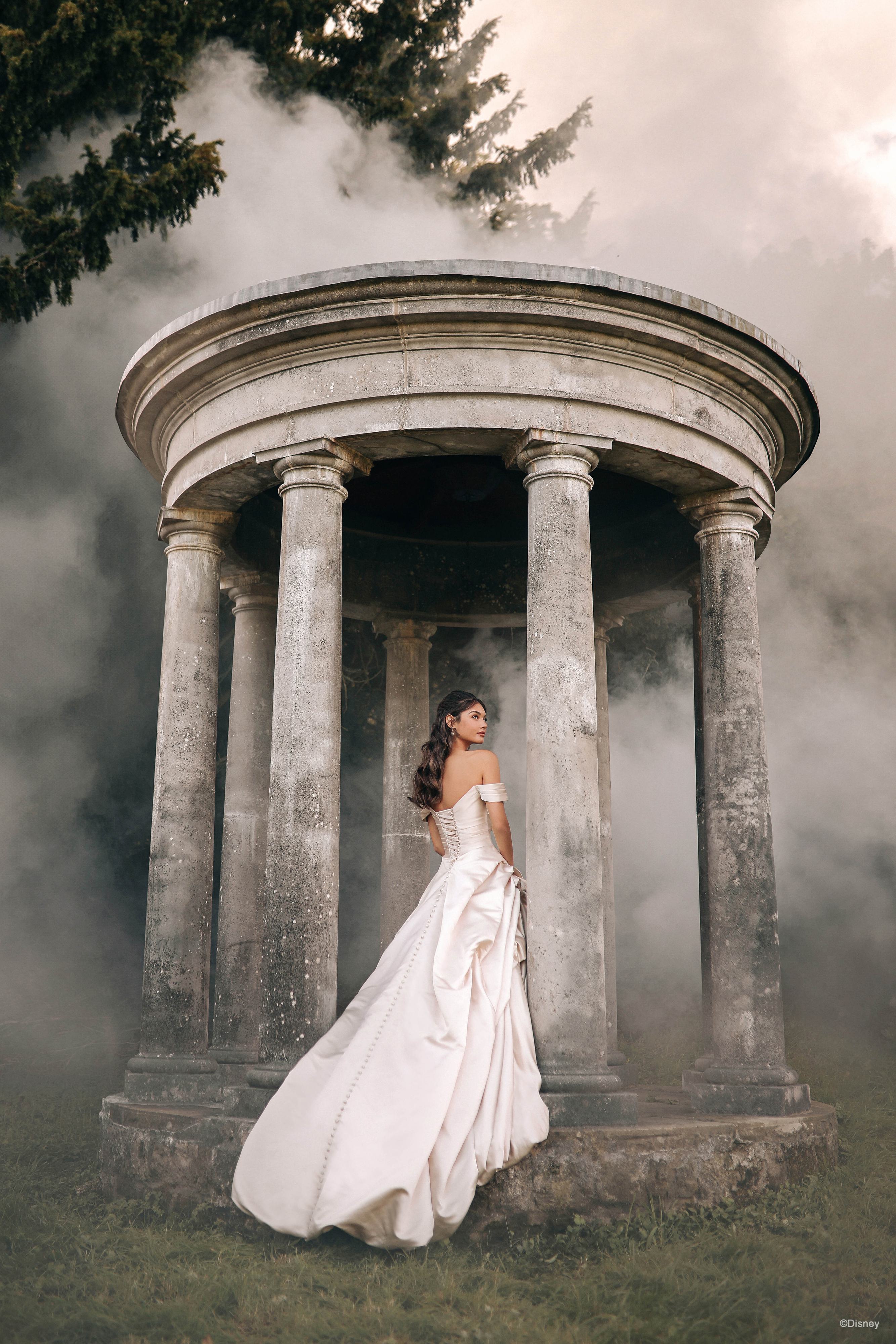 The image shows a woman wearing a luxurious and dramatic white wedding gown. This image is a view from the back of the dress.The dress features an off-the-shoulder neckline and a fitted bodice that accentuates the waist. The skirt is exceptionally full and voluminous, with elaborate draping and folds that create a grand and opulent silhouette. The fabric appears to be heavy and rich, contributing to the gown's regal and elegant appearance. The long train flows behind her, adding to the overall majestic and sophisticated look of the dress. The woman's hair is styled in loose waves, enhancing the romantic and timeless quality of the ensemble.