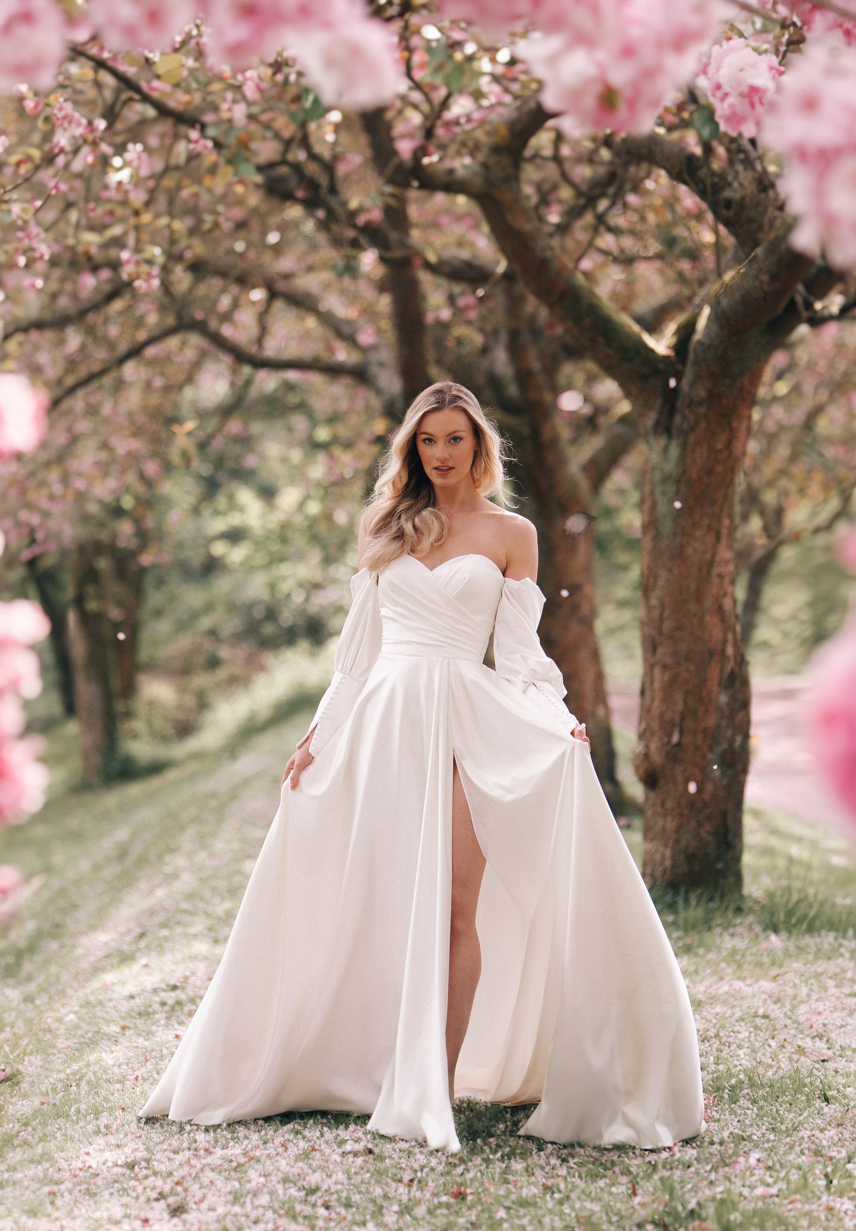 An elegant off-the-shoulder wedding dress with long, bishop sleeves and a sweetheart neckline, featuring a fitted bodice with pleated detailing and a flowing A-line skirt with a high slit for a touch of modern sophistication.