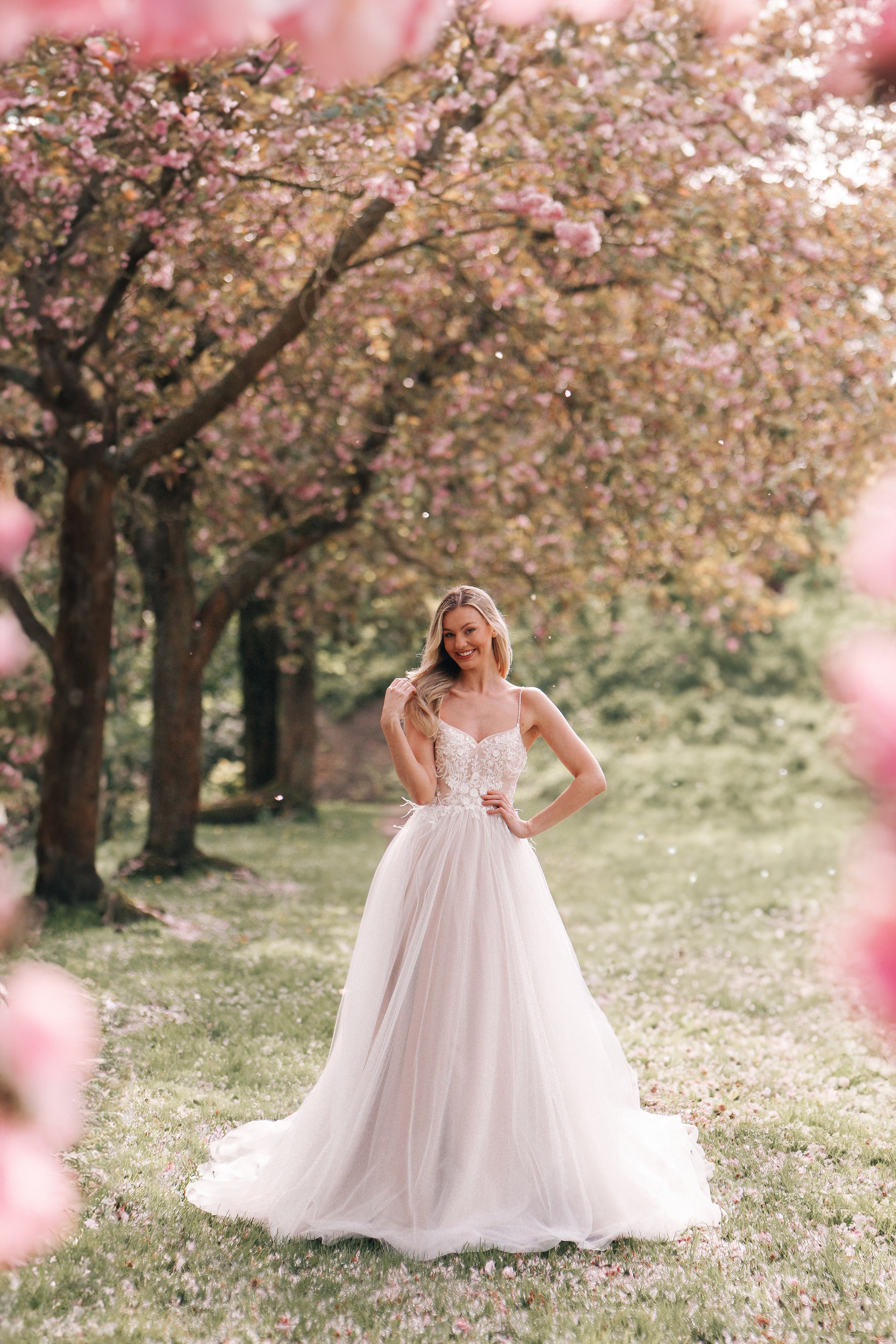 A bride stands amidst a picturesque garden filled with blooming cherry blossoms. She wears a stunning wedding gown featuring a fitted lace bodice with delicate spaghetti straps and a voluminous tulle skirt that flows gracefully to the ground. Her long blonde hair cascades over her shoulder as she gently holds a lock, with a serene smile gracing her face. The soft light filtering through the trees and the scattering petals create a magical, fairytale-like atmosphere, enhancing the beauty and elegance of the scene.