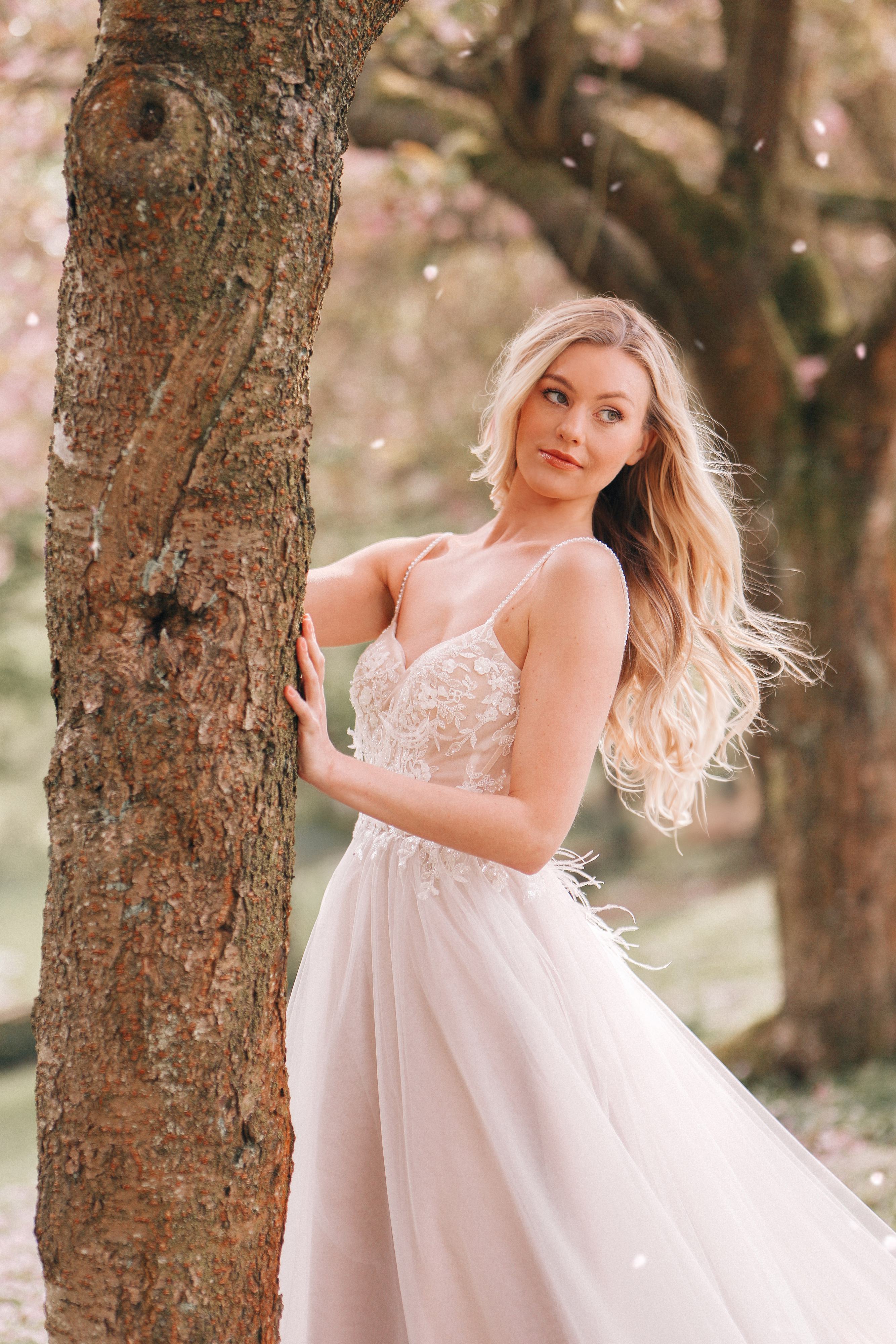 A bride stands gracefully beside a tree, gently holding onto its trunk. She is dressed in a delicate, flowing wedding gown with a lace bodice and thin straps. Her long, blonde hair cascades in soft waves over her shoulder. The background features a serene and natural setting with soft, pink blossoms and a gentle, magical atmosphere. The bride's expression is serene and reflective, adding to the romantic and enchanting mood of the scene.