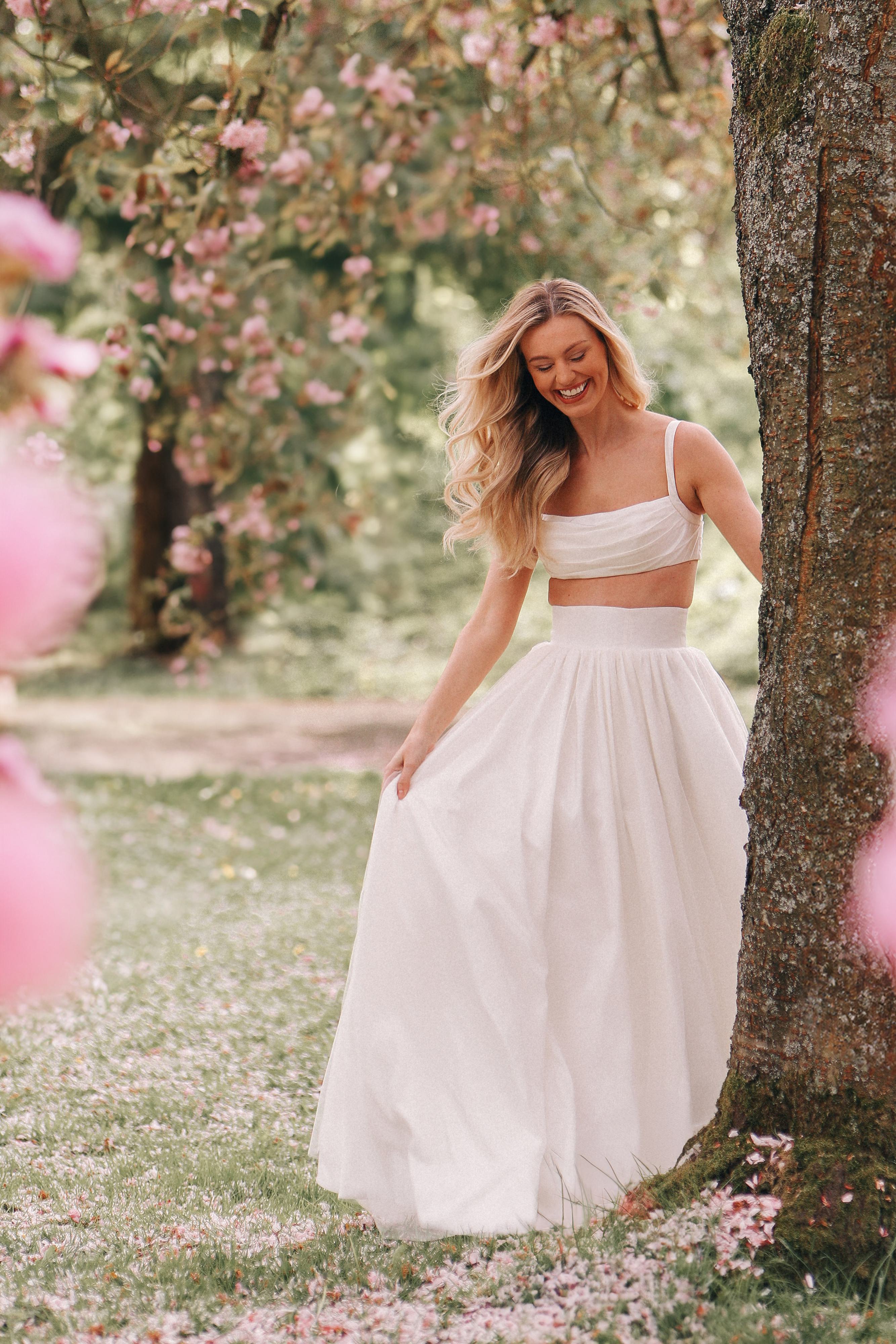 A joyful outdoor scene featuring a model in a bridal outfit by Lotte Nori Ivy. The dress is a two-piece design with a fitted, crop-top bodice and a full, flowing skirt. The model's long, blonde hair is styled in loose waves, and she is smiling brightly as she stands near a tree. The background is a picturesque garden filled with blooming pink flowers and lush greenery, adding to the romantic and whimsical atmosphere of the photo. The model's pose and the natural setting create a sense of carefree elegance and beauty.