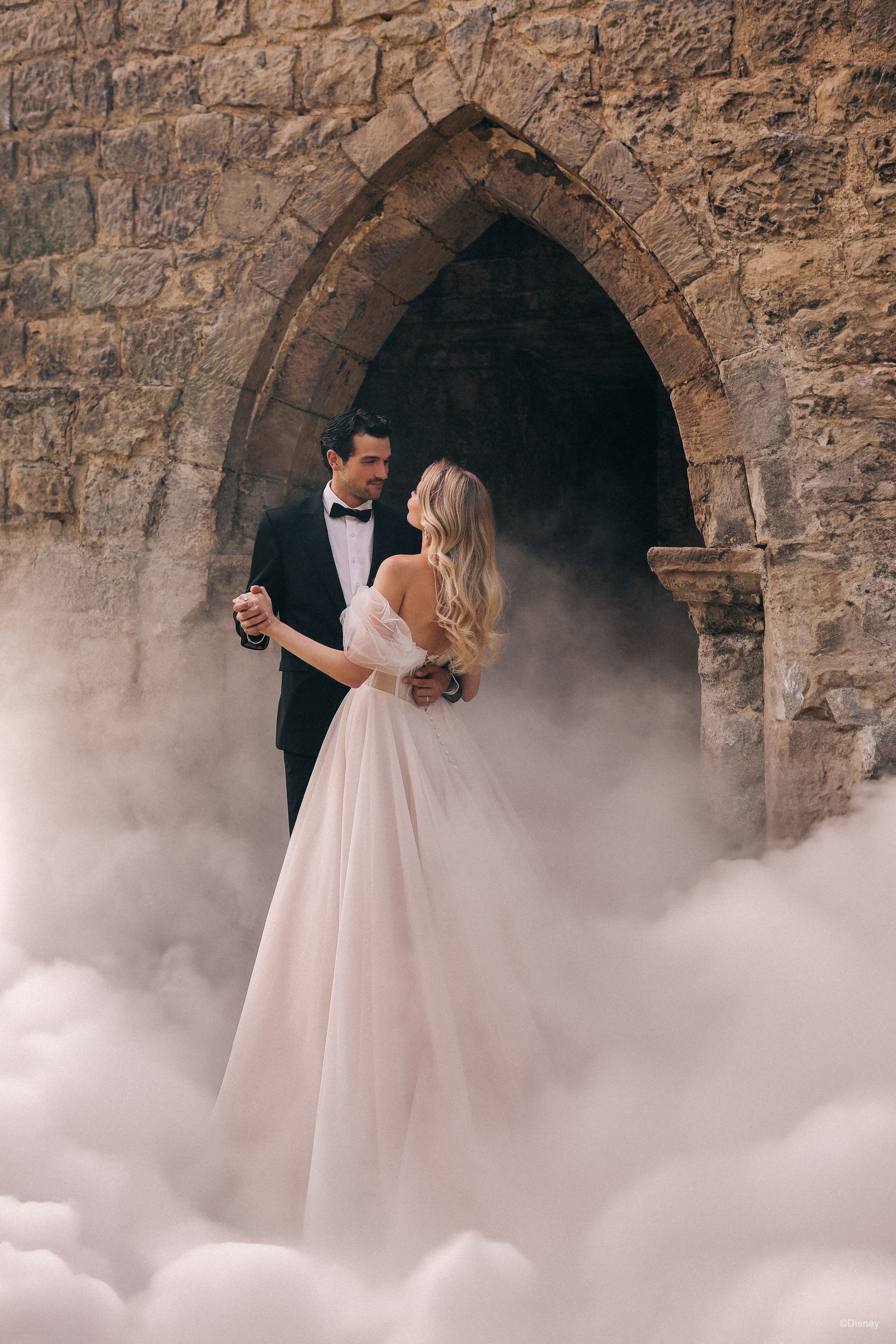 The image shows a couple dancing. The woman is wearing a white wedding gown with off-the-shoulder sleeves and her long blonde hair is styled in loose waves cascading down her back. The man is dressed in a classic black tuxedo with a white shirt and black bow tie. The couple appears to be sharing a romantic moment, gazing into each other's eyes as they dance, exuding a sense of joy and intimacy.
