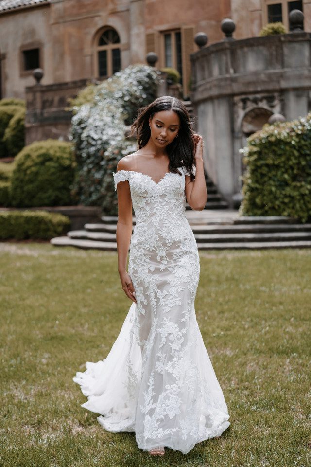An off-the-shoulder lace wedding gown featuring intricate floral appliqués throughout the fitted bodice and skirt. The dress has a sweetheart neckline and a semi-sheer overlay, adding a touch of allure. The lace detail extends down the mermaid silhouette, flaring out subtly at the knees and leading into a delicate train. The gown combines classic elegance with modern sophistication.
