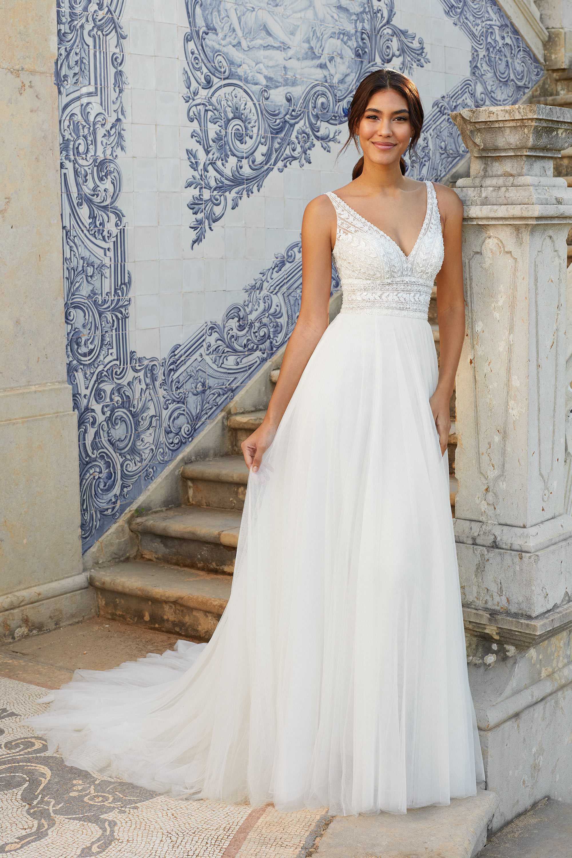 A bride stands in front of intricate blue and white tiled walls, wearing a Sincerity Bridal gown. The dress features a deep V-neckline, with a beaded bodice and delicate lace details that accentuate her waist. The flowing tulle skirt cascades gracefully to the ground, creating an ethereal silhouette. The bride’s hair is styled in a loose updo, enhancing the romantic and timeless look of the dress. The stone steps and ornate architectural backdrop add a touch of old-world charm to the scene.