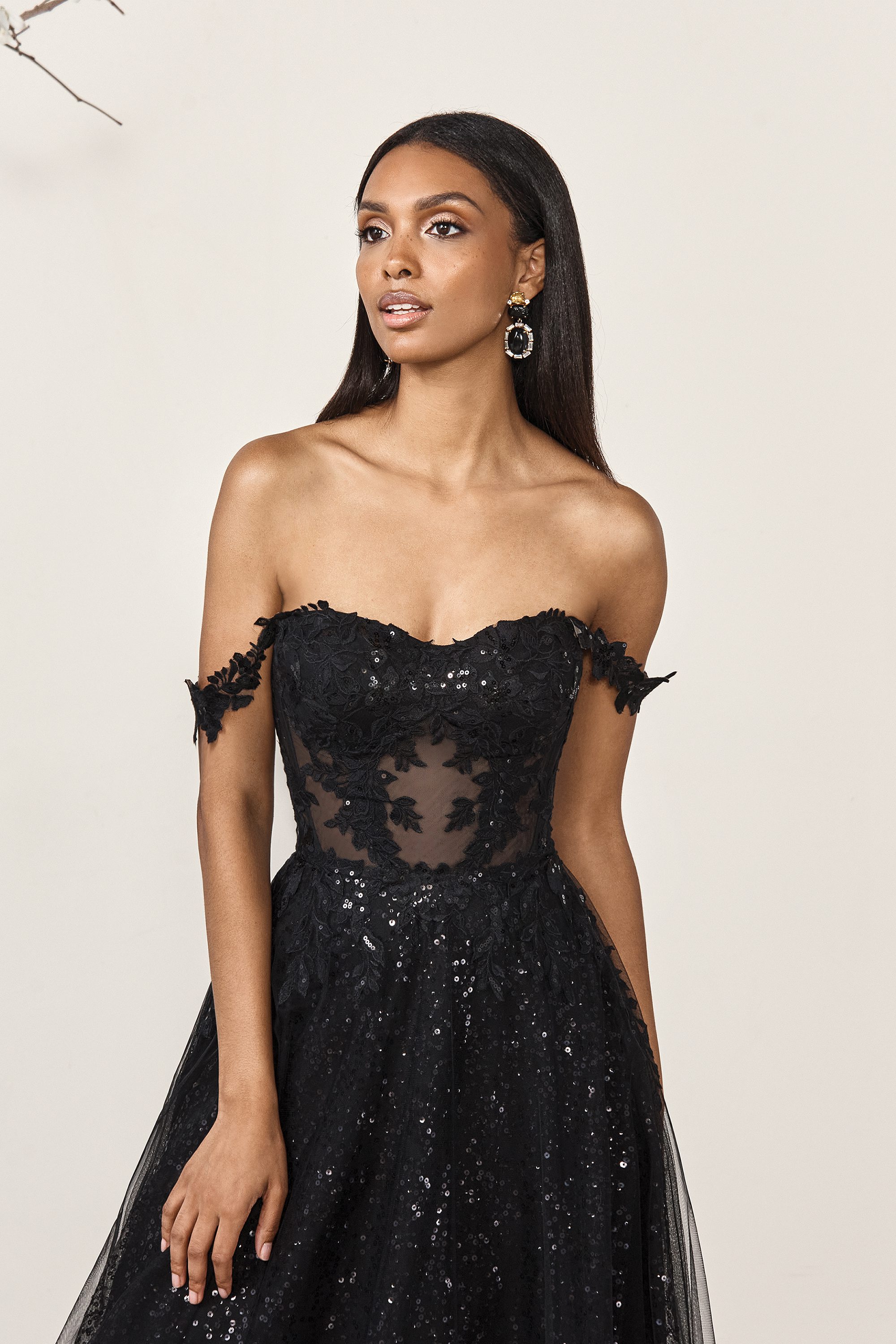 The bride wears a stunning black gown by Sincerity Bridal, featuring a sheer, off-the-shoulder bodice adorned with intricate floral lace appliqués. The fitted bodice elegantly contrasts with the voluminous skirt, which sparkles with subtle sequins, adding a touch of glamour. Her sleek, straight hair frames her face, highlighting her delicate features, while her confident expression complements the bold and sophisticated style of the gown. The overall look exudes modern elegance with a daring twist, perfect for a bride who wants to make a statement.