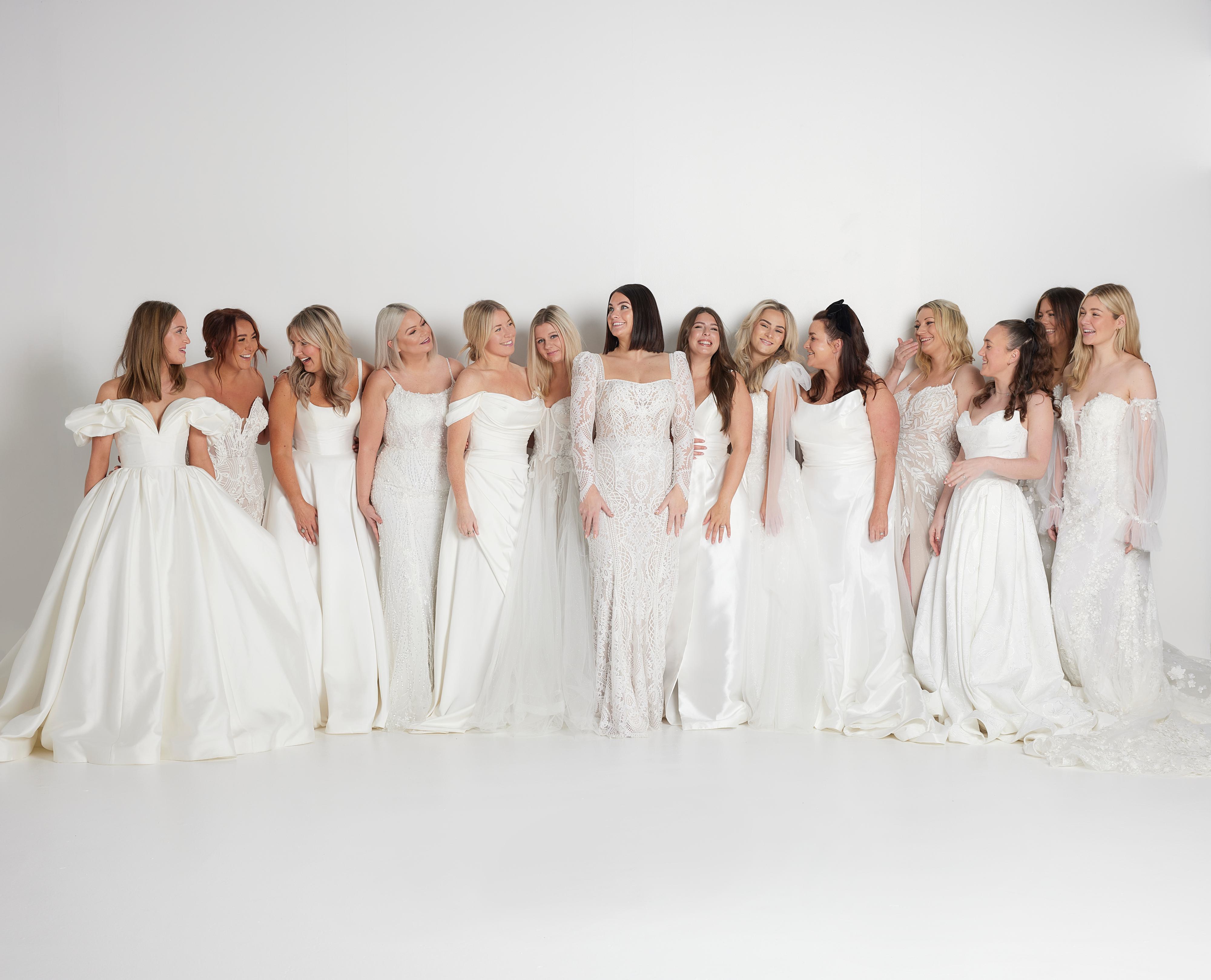 The image features a group of six women standing closely together, all wearing white bridal gowns. Each gown has a unique design, showcasing different styles and details. The women are smiling and appear to be enjoying the moment, with some leaning into each other in a friendly and supportive manner.
From left to right:
The first woman has long blonde hair and is wearing an off-the-shoulder dress.
The second woman has shoulder-length blonde hair and is also wearing an off-the-shoulder dress.
The third woman has blonde hair styled in loose waves and is wearing a strapless dress.
The fourth woman, positioned in the centre, has dark hair and is wearing a dress with sheer, lace-detailed sleeves.
The fifth woman has long dark hair and is wearing a deep V-neck dress with thin straps.
The sixth woman has long blonde hair and is wearing a dress with cap sleeves and a V-neckline.
The background is a plain, light colour, keeping the focus on the women and their elegant dresses. The overall mood of the image is joyful and celebratory, capturing the essence of bridal camaraderie and elegance.