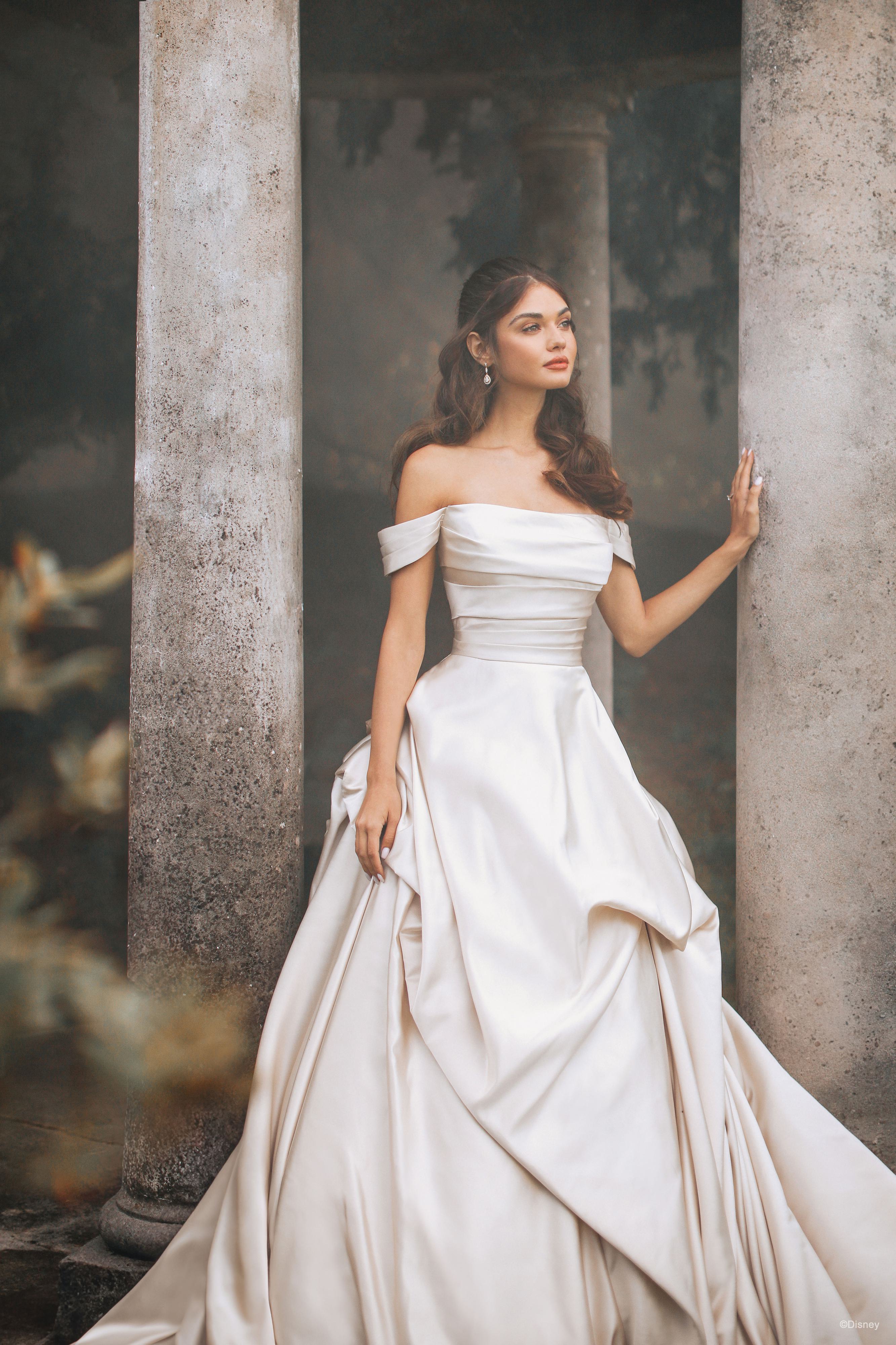 The image shows a woman wearing a luxurious and dramatic white wedding gown. The dress features an off-the-shoulder neckline and a fitted bodice that accentuates the waist. The skirt is exceptionally full and voluminous, with elaborate draping and folds that create a grand and opulent silhouette. The fabric appears to be heavy and rich, contributing to the gown's regal and elegant appearance. The long train flows behind her, adding to the overall majestic and sophisticated look of the dress. The woman's hair is styled in loose waves, enhancing the romantic and timeless quality of the ensemble.