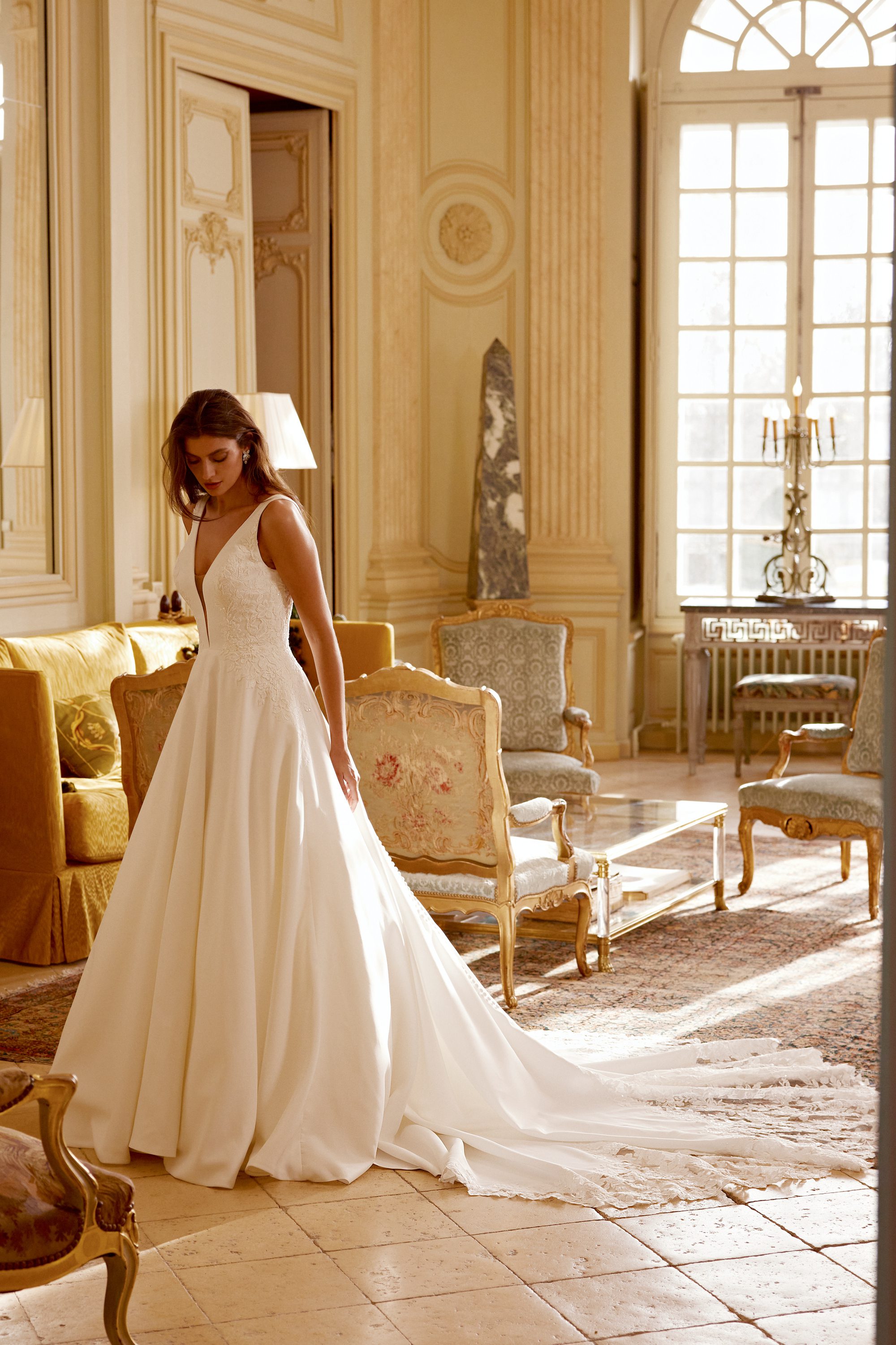 The image features the Gibson gown by Justin Alexander, set within an opulent and grand interior. The gown boasts a deep V-neckline and a fitted bodice that transitions into a voluminous skirt with a long, flowing train. The luxurious fabric catches the light beautifully, highlighting the intricate details of the gown. The model stands gracefully in a sunlit room adorned with classic furniture and elegant decor, including golden chairs and large windows that allow soft, natural light to illuminate the space. The overall ambiance is one of timeless elegance and sophistication, perfectly complementing the refined beauty of the Gibson gown.