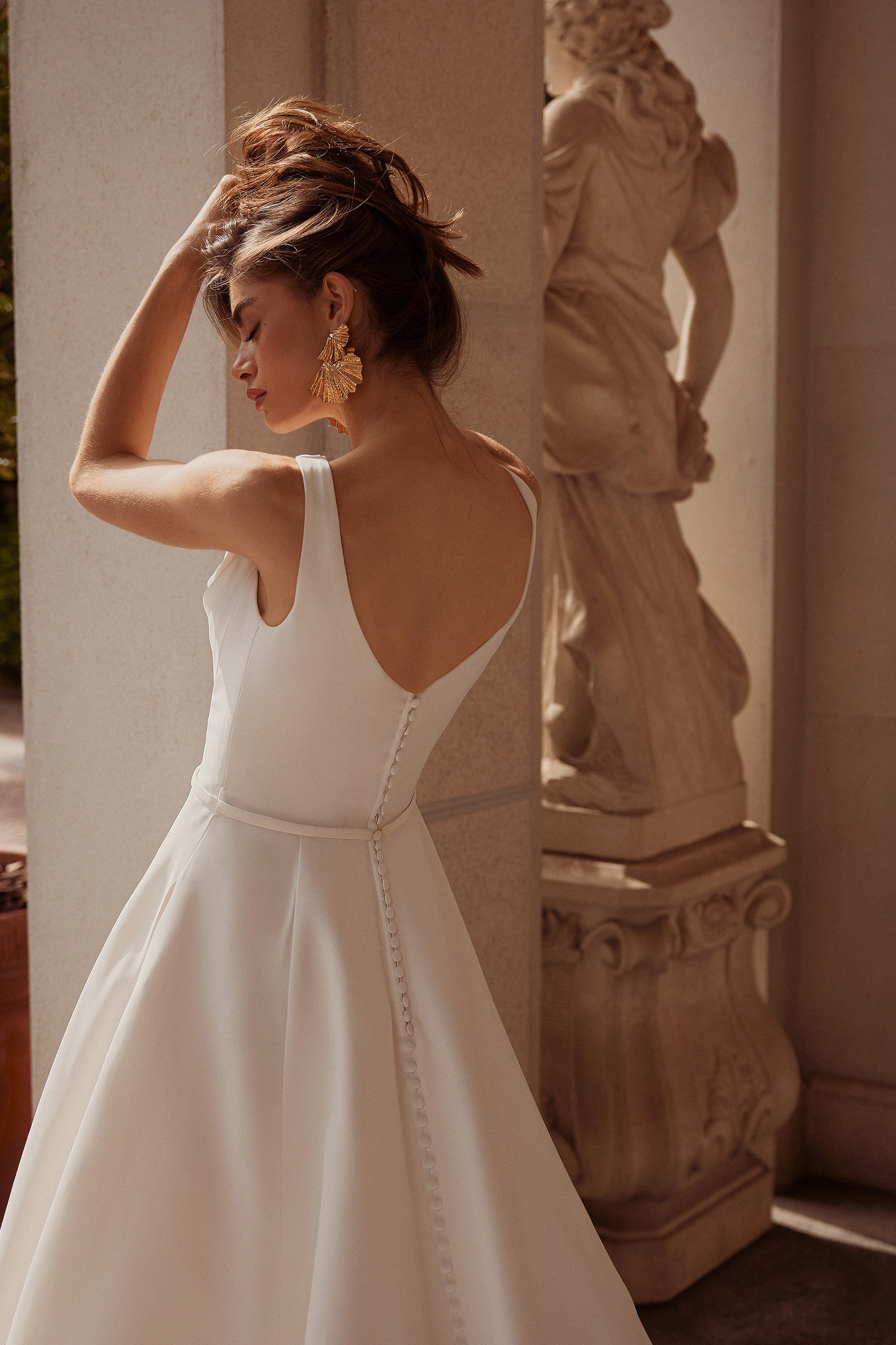 The image features the Grove gown by Justin Alexander, highlighting the back view of the dress in a sophisticated and serene setting. The model poses with one arm gracefully lifting her hair, showcasing the deep V-back of the gown, which is adorned with a row of delicate buttons that extend down to the waistline. The dress is tailored to perfection, with the fabric flowing smoothly into a full A-line skirt. The simplicity of the design is elevated by the model's choice of statement gold earrings, adding a touch of glamour. The background features classical architecture, with a stone statue partially visible, enhancing the gown's timeless elegance.