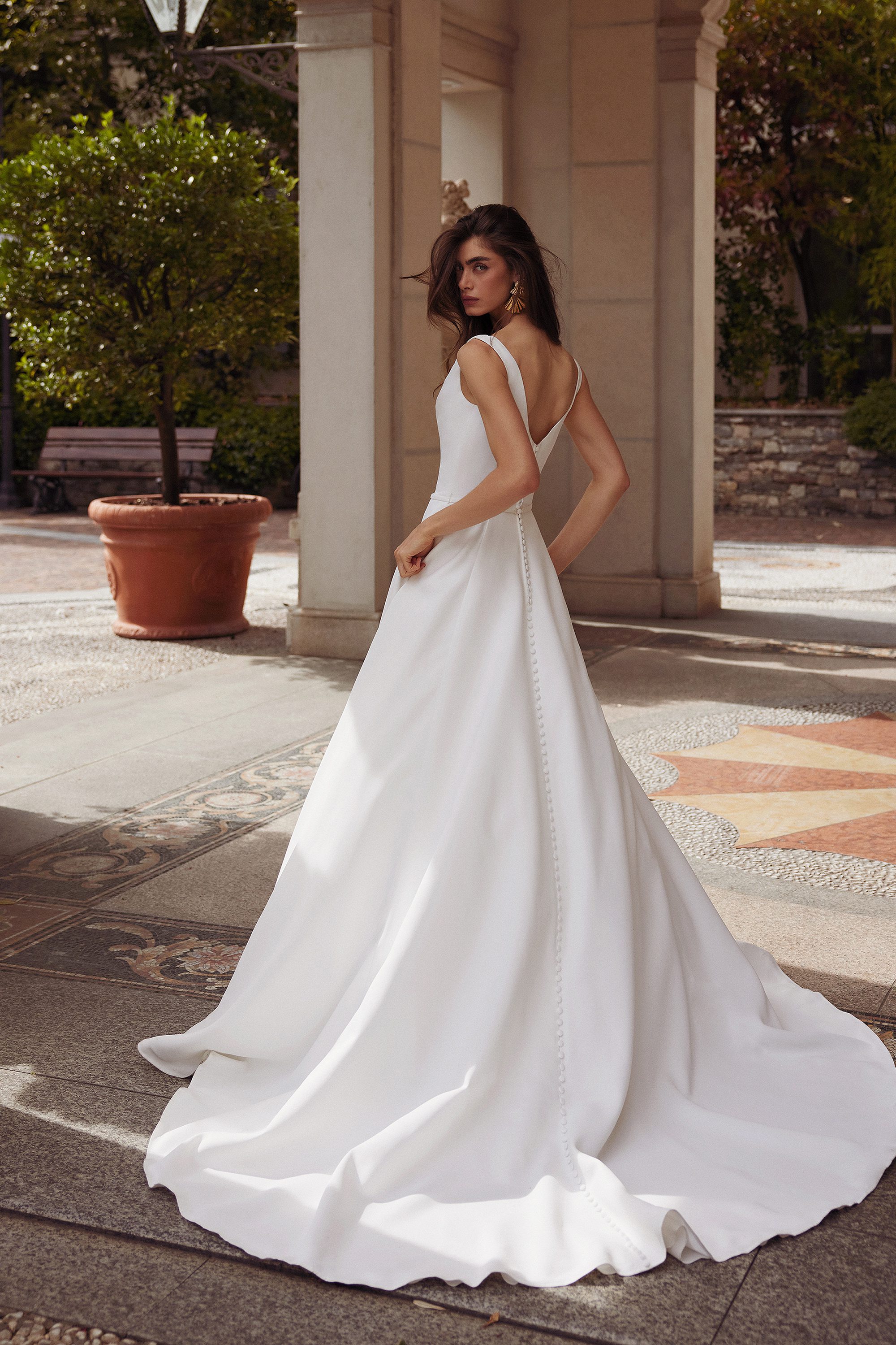 The image shows the Grove gown by Justin Alexander, captured from the back as the model turns slightly to reveal the dress’s flowing A-line silhouette. The gown features a deep scoop back that adds a touch of allure to the otherwise modest design. A row of delicate buttons runs down the center of the dress, from the back of the bodice to the train, emphasizing the gown’s refined craftsmanship. The fabric drapes elegantly to the floor, creating a subtle yet dramatic train that glides over the patterned stone surface beneath. The outdoor setting, with its classical architecture and greenery, complements the gown's timeless elegance.