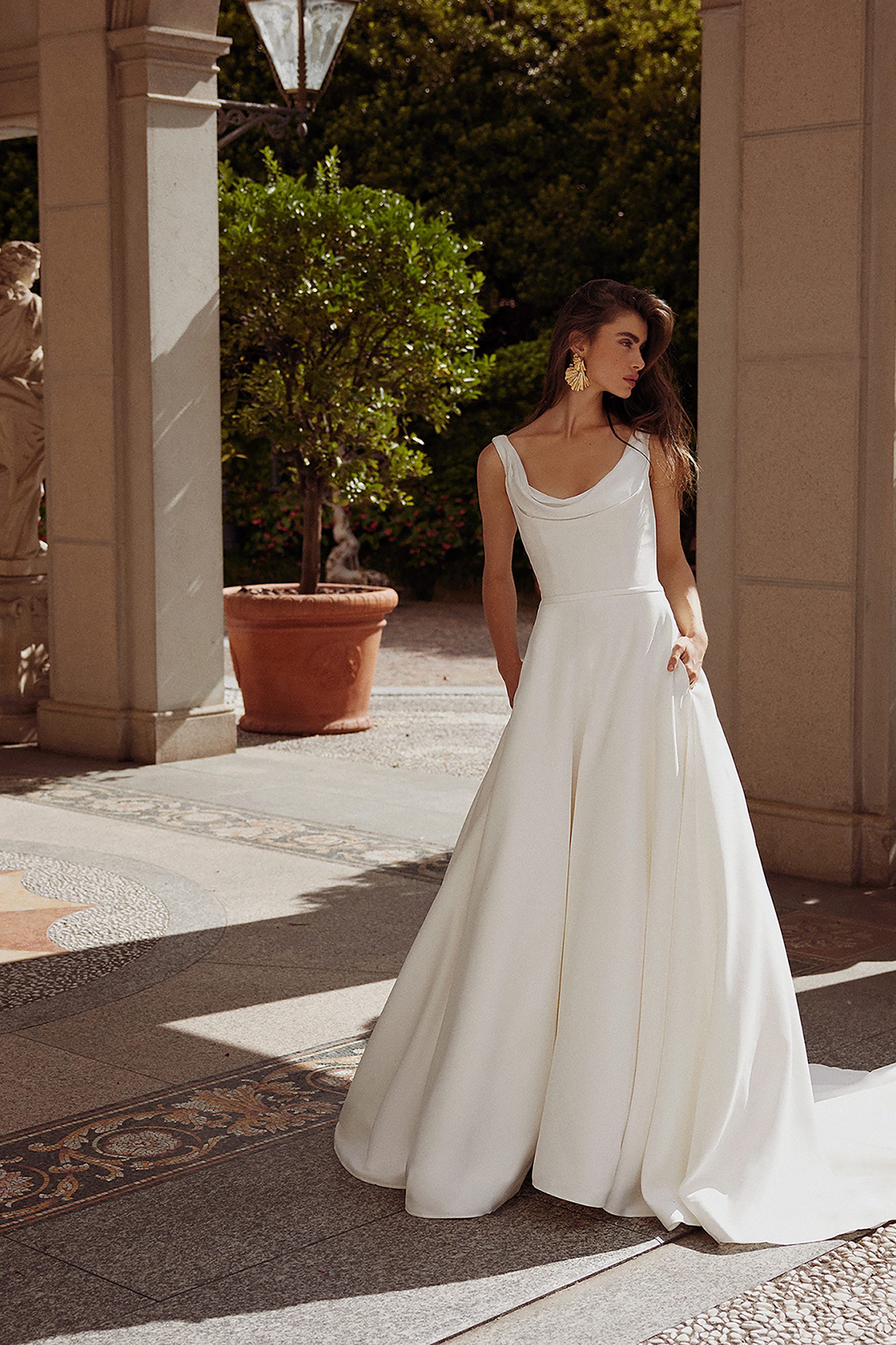 The image showcases the Grove gown by Justin Alexander, captured in an elegant outdoor setting. The model wears a floor-length A-line dress with a soft cowl neckline that gracefully drapes across the bodice, creating a romantic and timeless look. The wide straps add a touch of modernity while ensuring comfort and support. The dress features hidden pockets, which the model casually rests her hands in, adding a chic and relaxed vibe to the otherwise classic design. The gown's fabric flows beautifully to the ground, creating gentle folds that enhance its voluminous skirt. The backdrop, with stone columns and lush greenery, complements the gown’s sophisticated and serene aesthetic.