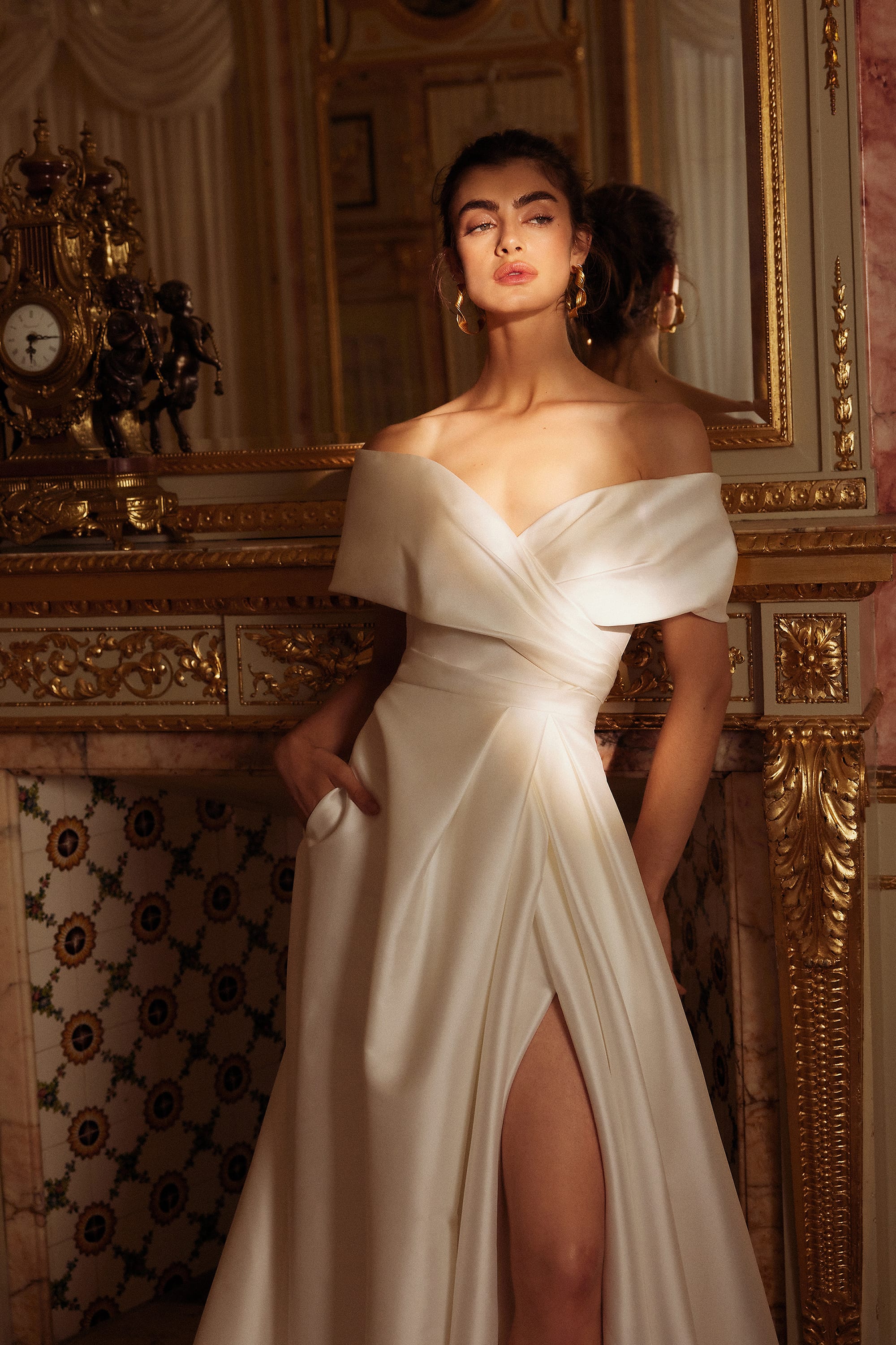 A woman is standing in an opulent, classical room, dressed in an elegant off-the-shoulder white gown. The gown has a fitted bodice with a wrapped design and a high slit on the left side, revealing part of her leg. The fabric flows gracefully to the floor, forming a small train behind her. She is standing in front of an ornate fireplace, adorned with intricate gold details and a large mirror above it. The room features rich decorations, including a vintage clock on the mantel, luxurious drapes, and detailed moldings. The overall ambiance is luxurious and sophisticated.