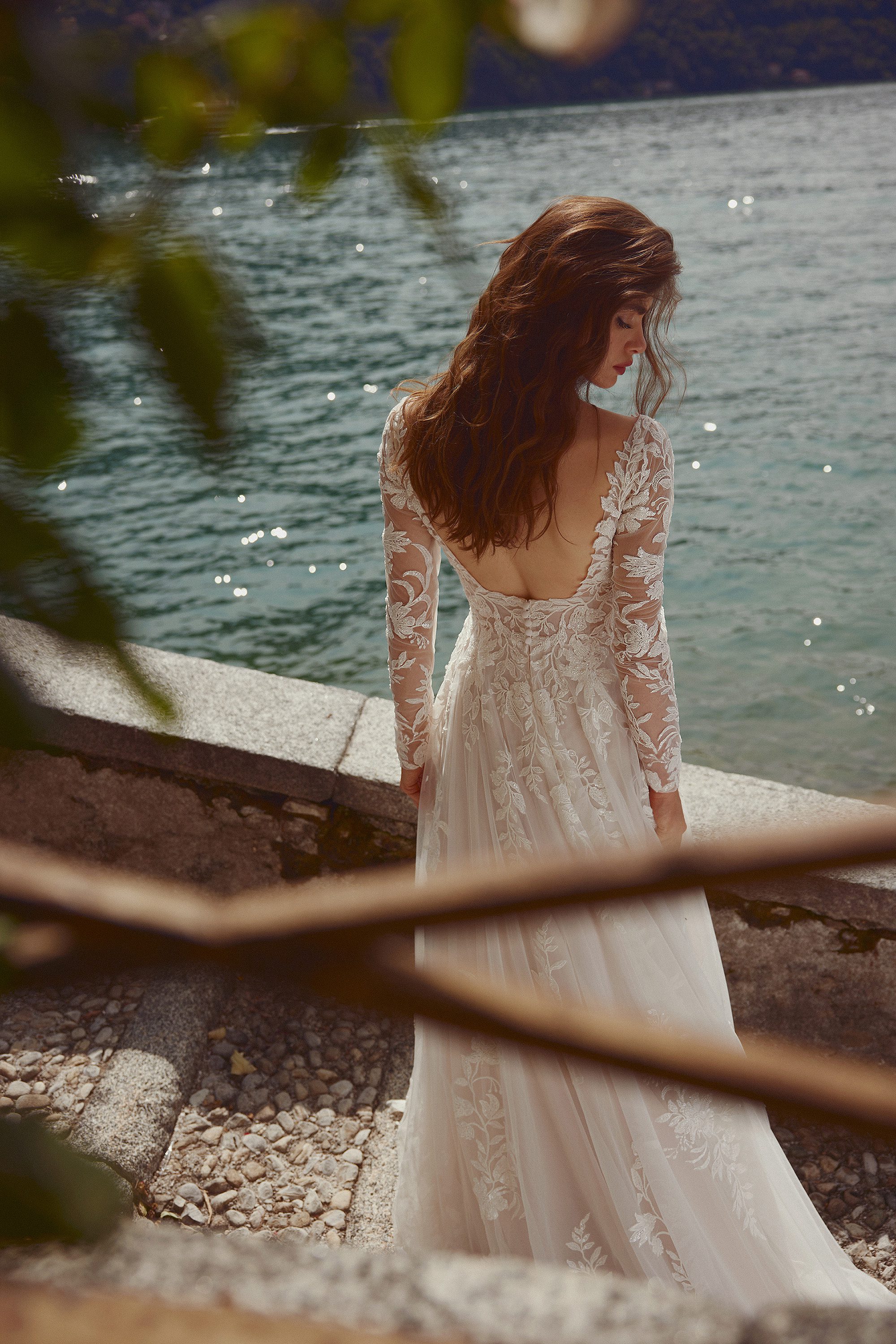 A bride in the Isley gown by Justin Alexander is captured in a moment of serene reflection by the water's edge. The gown's stunning open back and long, sheer lace sleeves are beautifully showcased as the bride gazes down, her loose waves gently falling over her shoulders. The intricate floral lace detailing of the dress contrasts elegantly with the soft ripples of the lake behind her. The scene is framed by blurred branches, creating an intimate, almost dreamlike atmosphere, as the sun's rays dance upon the water, highlighting the romantic and ethereal quality of the gown.