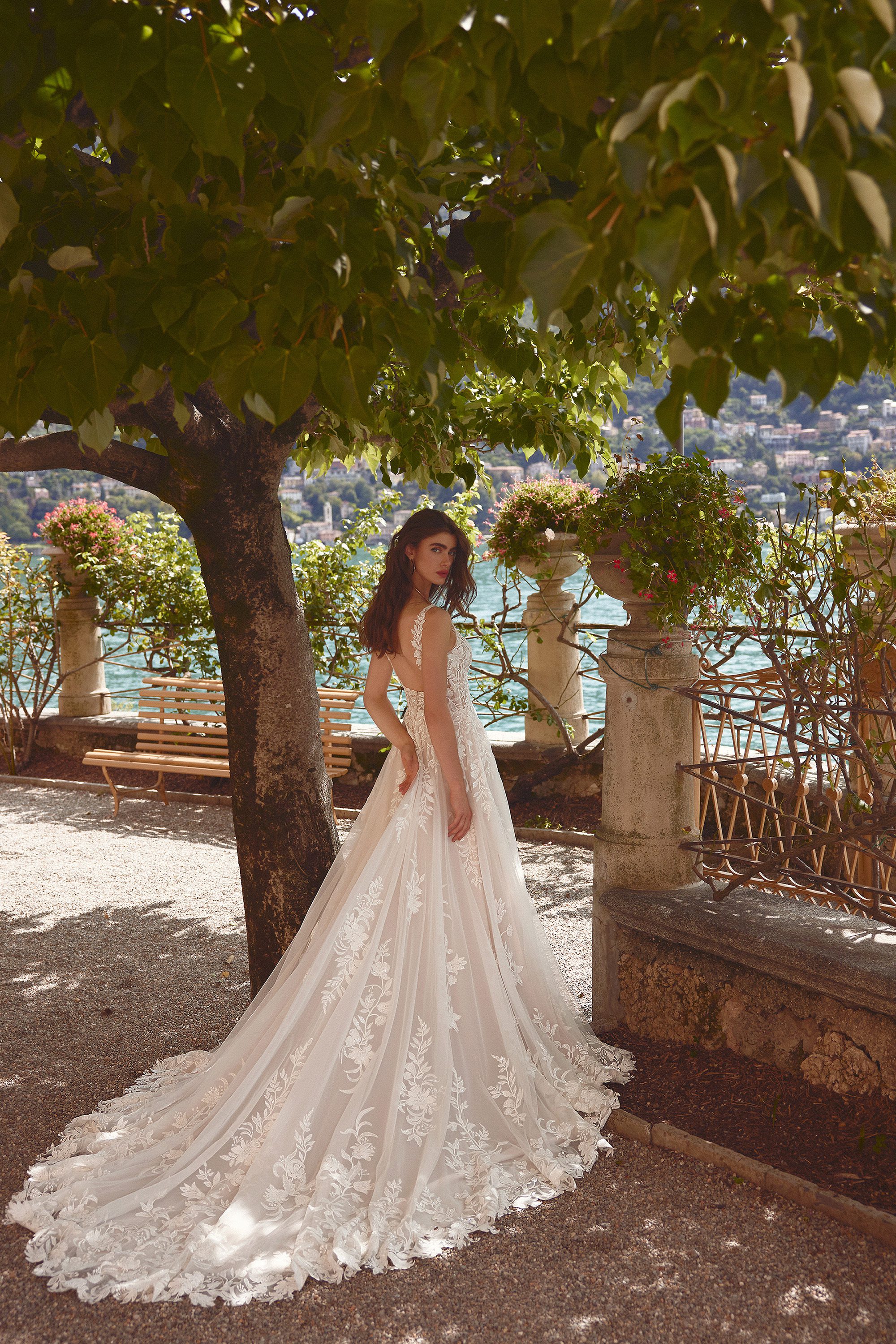 A bride in the Isley gown by Justin Alexander stands beneath the shade of a large tree, surrounded by the tranquil beauty of a lakeside garden. The dress, with its intricate lace details and plunging open back, drapes elegantly down her figure, culminating in a full, flowing train that sweeps across the ground. The delicate lace appliqués blend seamlessly with the natural setting, as the light filters through the leaves above, casting a soft glow over the scene. Stone pillars and a distant view of the shimmering lake create a serene and romantic backdrop, enhancing the timeless elegance of the gown.
