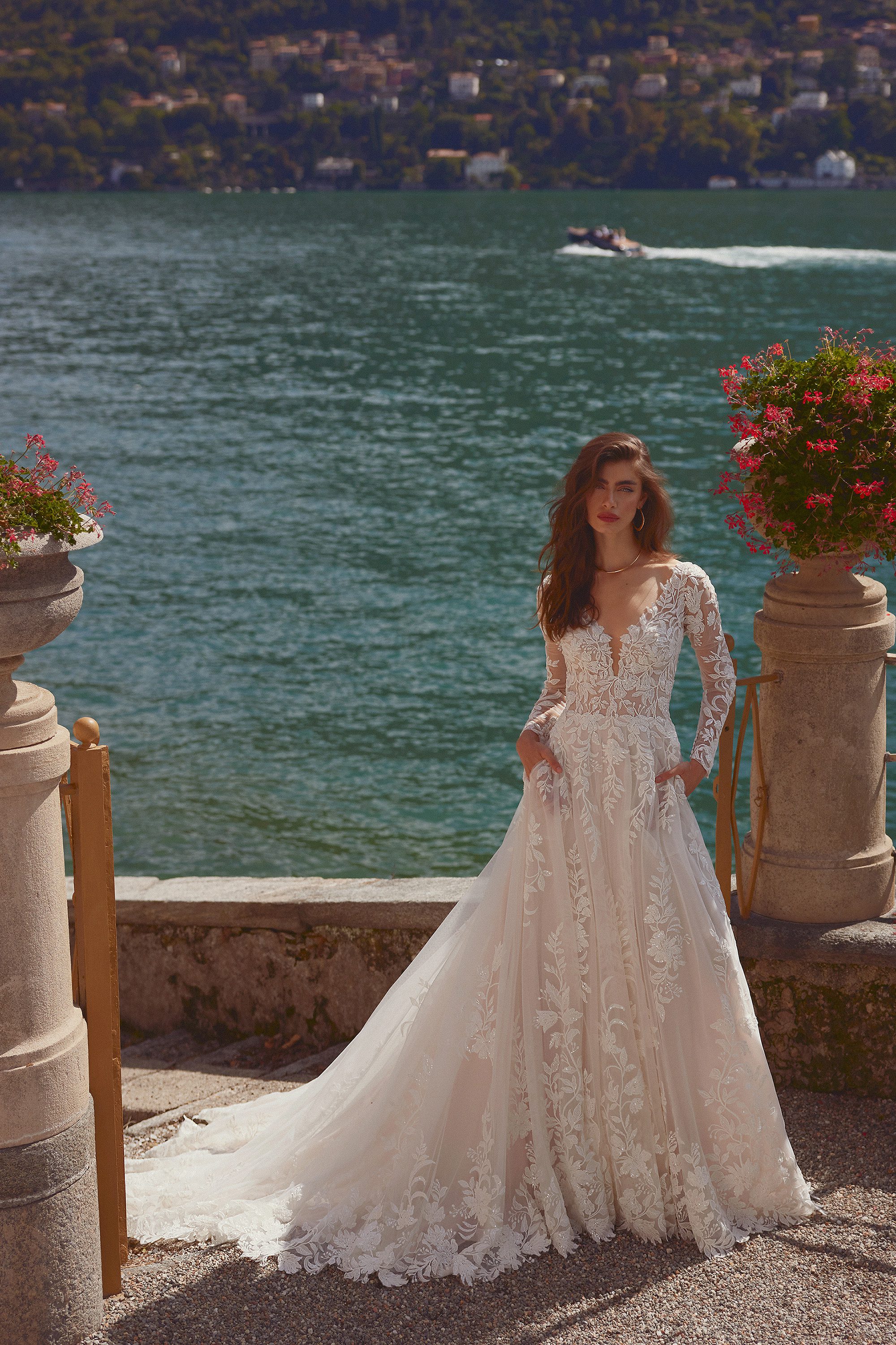 A bride stands gracefully by the edge of a serene lake, framed by ornate stone pillars adorned with vibrant flowers. She is wearing the Isley gown by Justin Alexander, a breathtaking lace wedding dress that features a deep V-neckline and long, sheer sleeves. The intricate lace appliqués cascade down the fitted bodice and flow into the full, voluminous skirt, which extends into a long, flowing train that gently sweeps the ground. The backdrop of the glistening lake and distant hills enhances the romantic elegance of the gown, creating a picture-perfect moment.