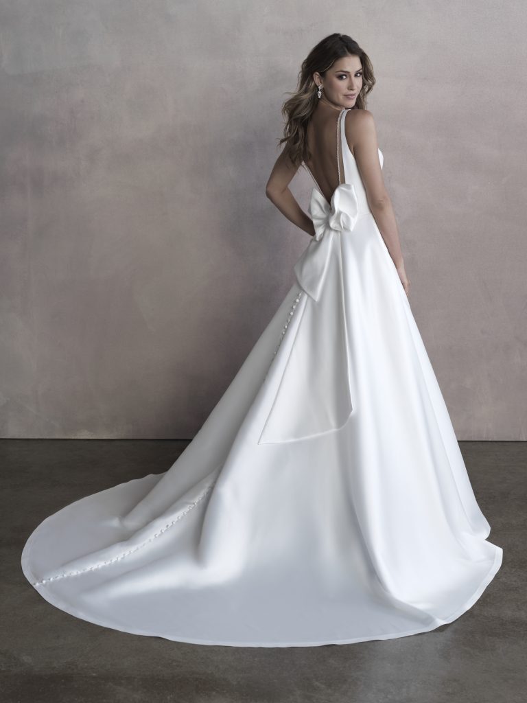 A bride is elegantly posed in a sleek, minimalist wedding gown with a deep V-back, revealing her back while a large, statement bow sits at the waist, adding a modern touch to the classic silhouette. The gown features a long, flowing train with a row of delicate buttons trailing down, enhancing its timeless beauty. Her hair is styled in soft waves, and she looks confidently over her shoulder, exuding grace and sophistication against a muted backdrop that emphasizes the simplicity and elegance of her attire.