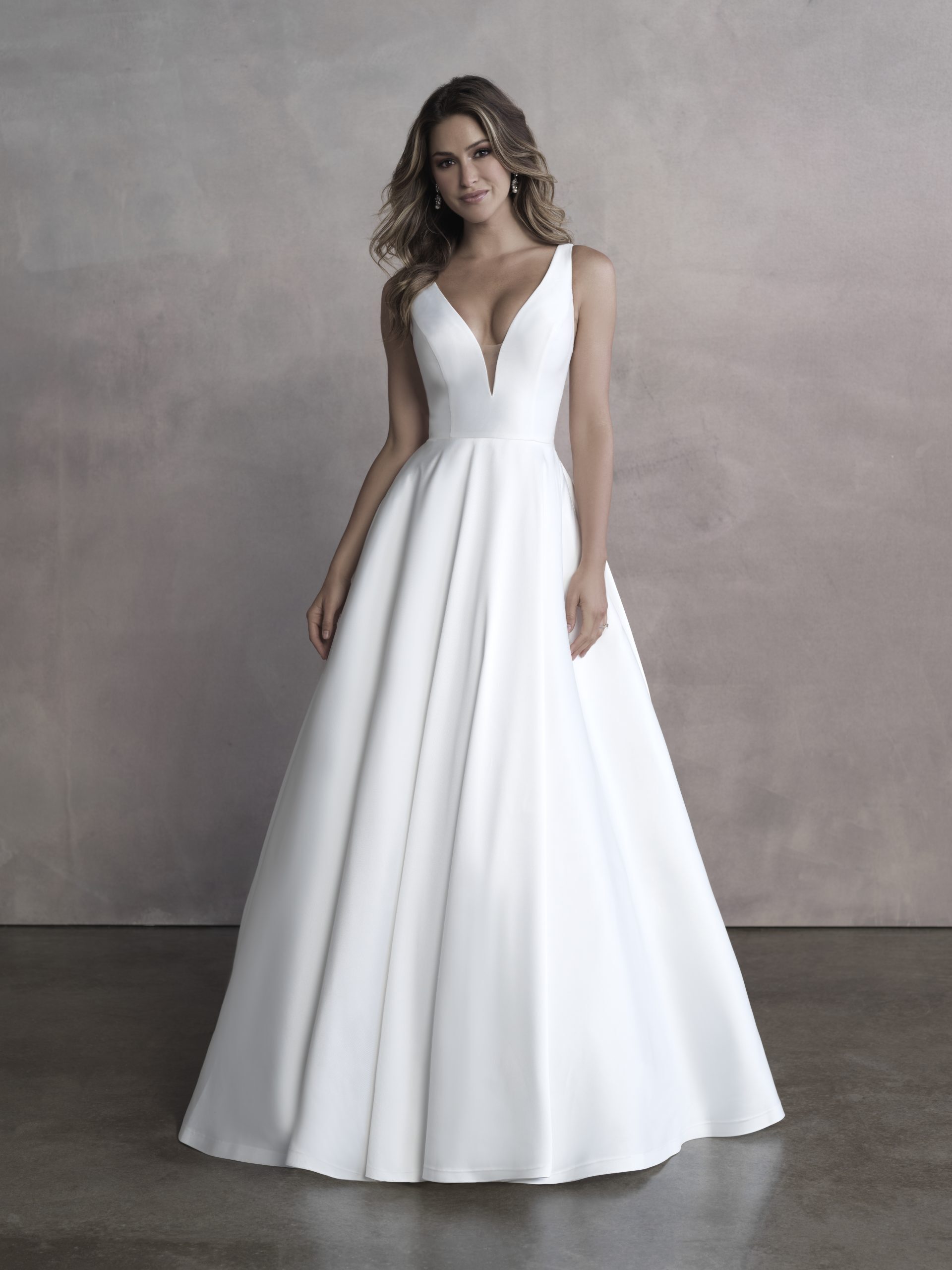 A bride stands confidently in a minimalist, modern wedding gown featuring a sleek, A-line silhouette. The dress is crafted from smooth, luxurious fabric that falls elegantly to the floor, creating a clean and sophisticated look. The bodice is fitted with a plunging V-neckline, adding a touch of allure while maintaining a timeless elegance. The gown has wide shoulder straps that frame the neckline and emphasize the bride's natural beauty. The backdrop is neutral, allowing the simplicity and beauty of the dress to take center stage.