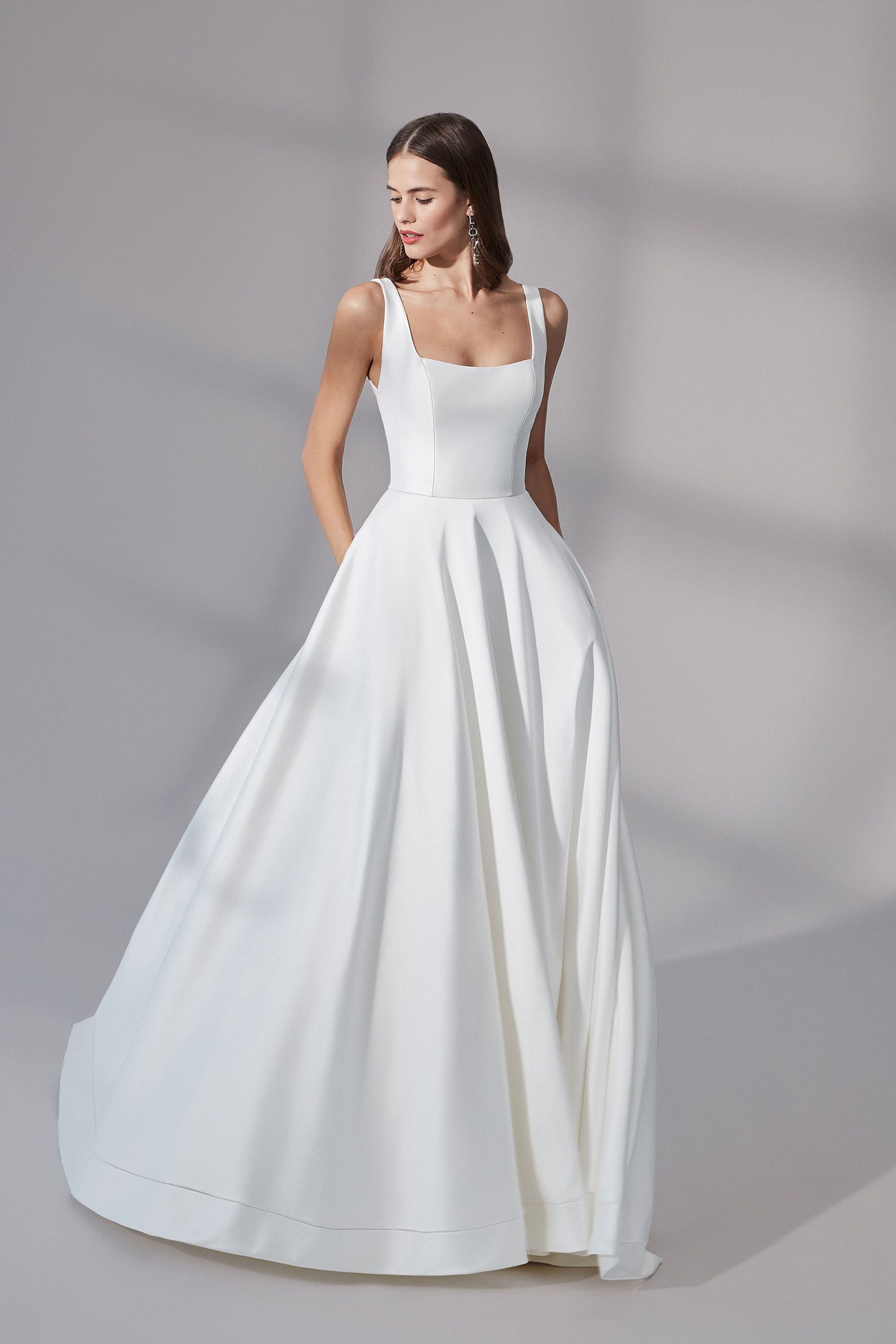 A bride in the Charleston gown by Justin Alexander, showcasing a classic A-line silhouette with a structured bodice and square neckline. The dress features wide straps and a flowing skirt with soft pleats, creating an elegant and timeless look. The clean, minimalist design highlights the luxurious fabric and impeccable tailoring, perfect for a modern bride who appreciates refined simplicity.