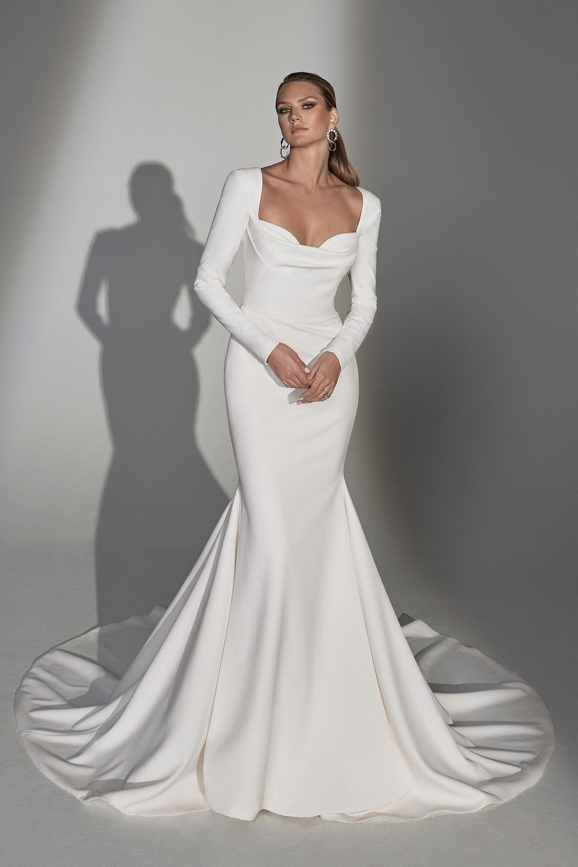 Front view of the Norma gown by Justin Alexander. This elegant wedding dress features a fitted silhouette with long sleeves and a dramatic, sweeping train. The bodice showcases a structured sweetheart neckline with a squared-off edge, offering a modern twist on a classic design. The smooth, sleek fabric of the gown emphasizes the bride's figure, while the extended train adds a touch of grandeur. The model's poised stance against a neutral background highlights the gown's timeless sophistication and refined detailing.