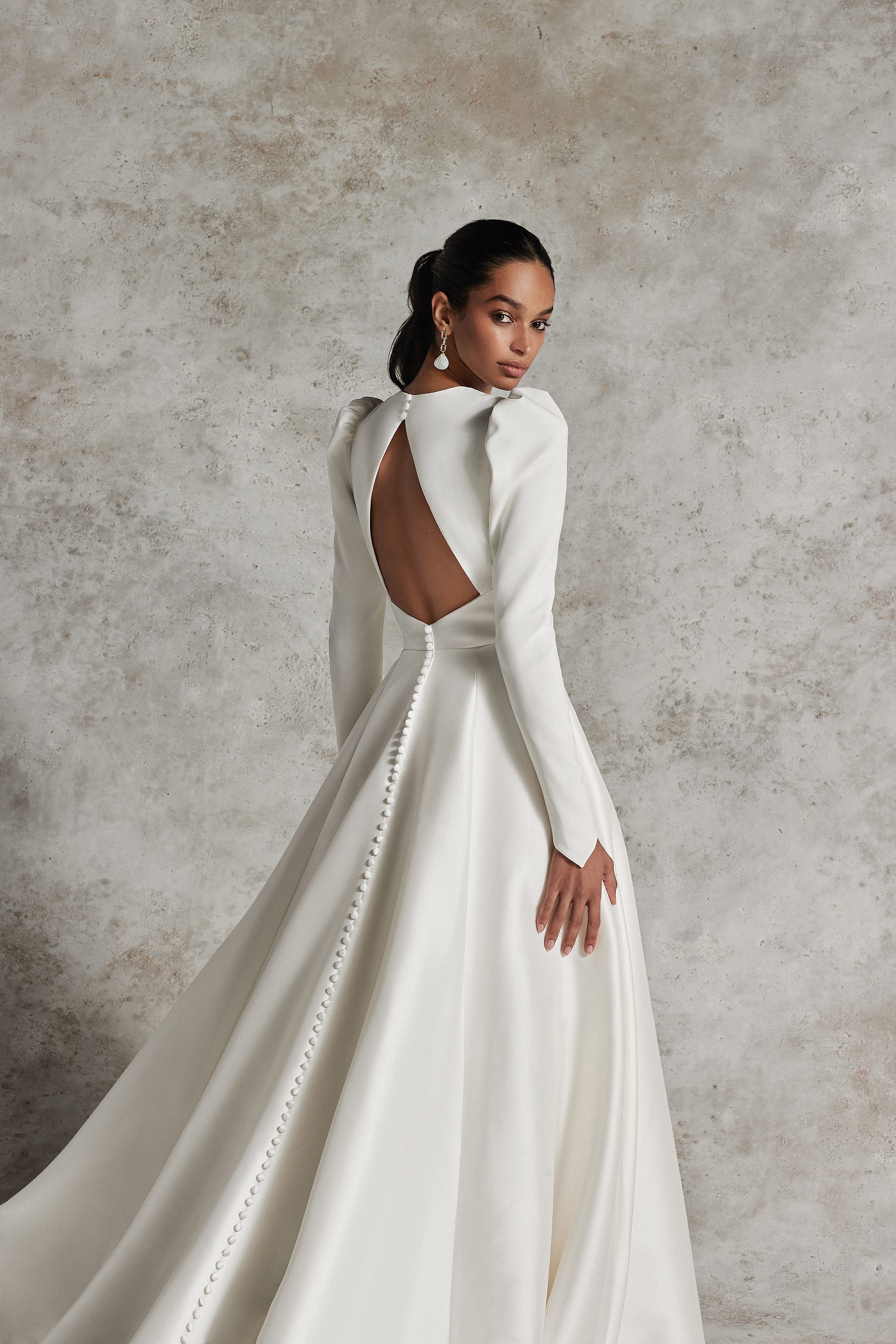 A bride models a stunning Justin Alexander dress, showcasing a sophisticated back design with a keyhole cutout. The dress features long sleeves and a full satin skirt adorned with a line of buttons trailing elegantly down the train.