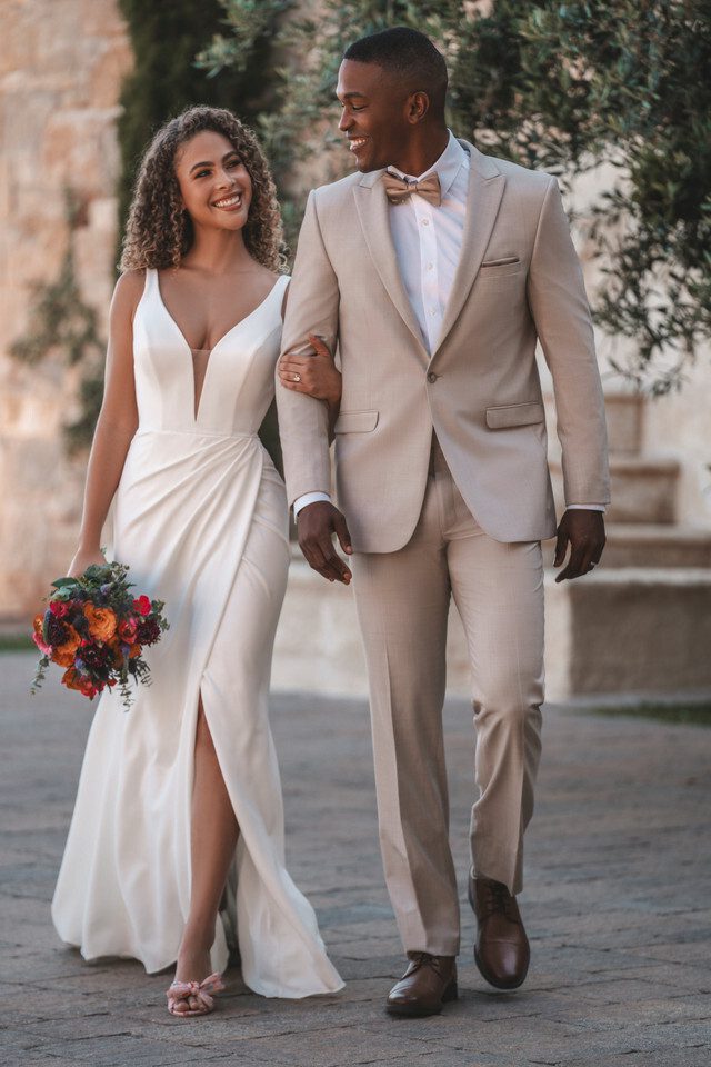 A joyful bride and groom walk arm in arm, exuding happiness and elegance. The bride wears a sleek, ivory gown with a deep V-neckline and a thigh-high slit, which adds a modern and chic touch to her bridal look. Her natural curls cascade around her shoulders, and she carries a vibrant bouquet of red, orange, and pink flowers. The groom complements her perfectly in a light beige suit paired with a white shirt and a stylish bow tie. The couple's smiles and the serene, sunlit surroundings with stone architecture and greenery create a warm, inviting atmosphere.