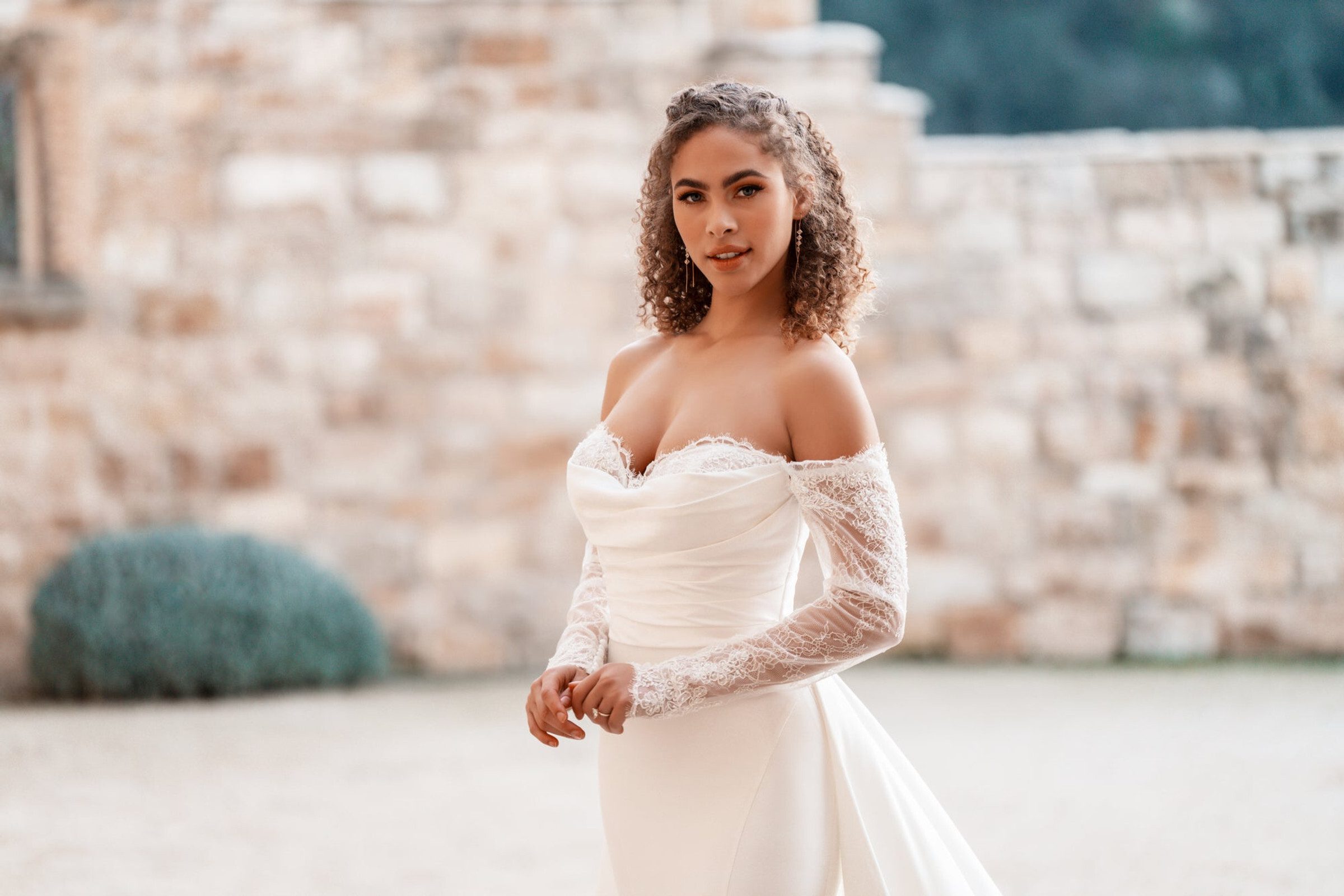This Allure Bridals dress features a captivating off-the-shoulder neckline adorned with delicate lace detailing. The fitted bodice is elegantly ruched, enhancing the bride's natural curves, while the long, sheer lace sleeves add a touch of romantic sophistication. The gown's silhouette flows seamlessly into a sleek skirt, creating a timeless and graceful look perfect for a classic wedding setting.