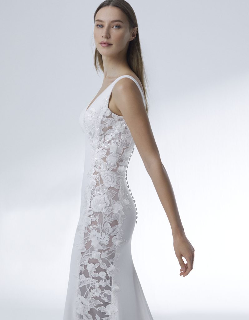 The image features the Alison wedding gown, highlighting its intricate lace side panels that run from the bodice down to the hips, creating a beautiful contrast against the smooth fabric of the dress. The gown is designed with a flattering fit that accentuates the bride's natural curves, with delicate buttons running along the side seam for added elegance. The shoulder straps provide a secure and comfortable fit, while the soft lace and floral appliqués offer a romantic touch. The model's poised expression complements the gown's refined and graceful aesthetic.