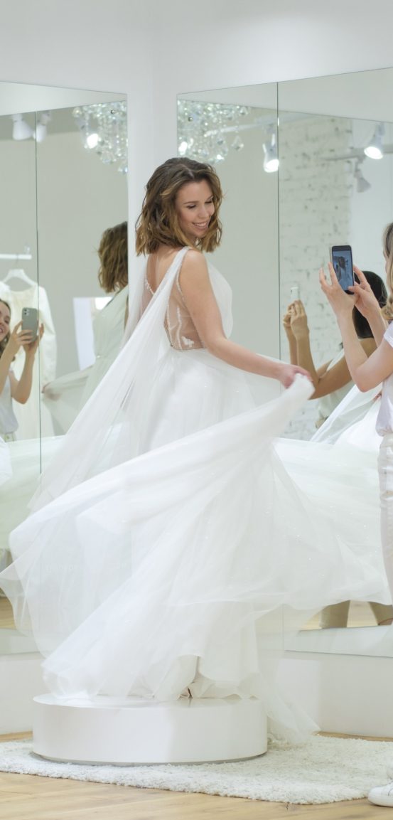 A joyful bride twirls gracefully on a raised platform in a bright, modern fitting room. She is wearing a voluminous white wedding gown that flows beautifully as she spins, capturing the movement of the fabric. The bride's expression is one of happiness, and her reflection is visible in the large mirrors surrounding her, adding depth to the image. Another person, likely a friend or consultant, stands nearby, capturing the moment on a smartphone. The room is sleek and well-lit, with minimalist decor that includes a chandelier and light-colored walls, creating a perfect backdrop for the bride's special moment.