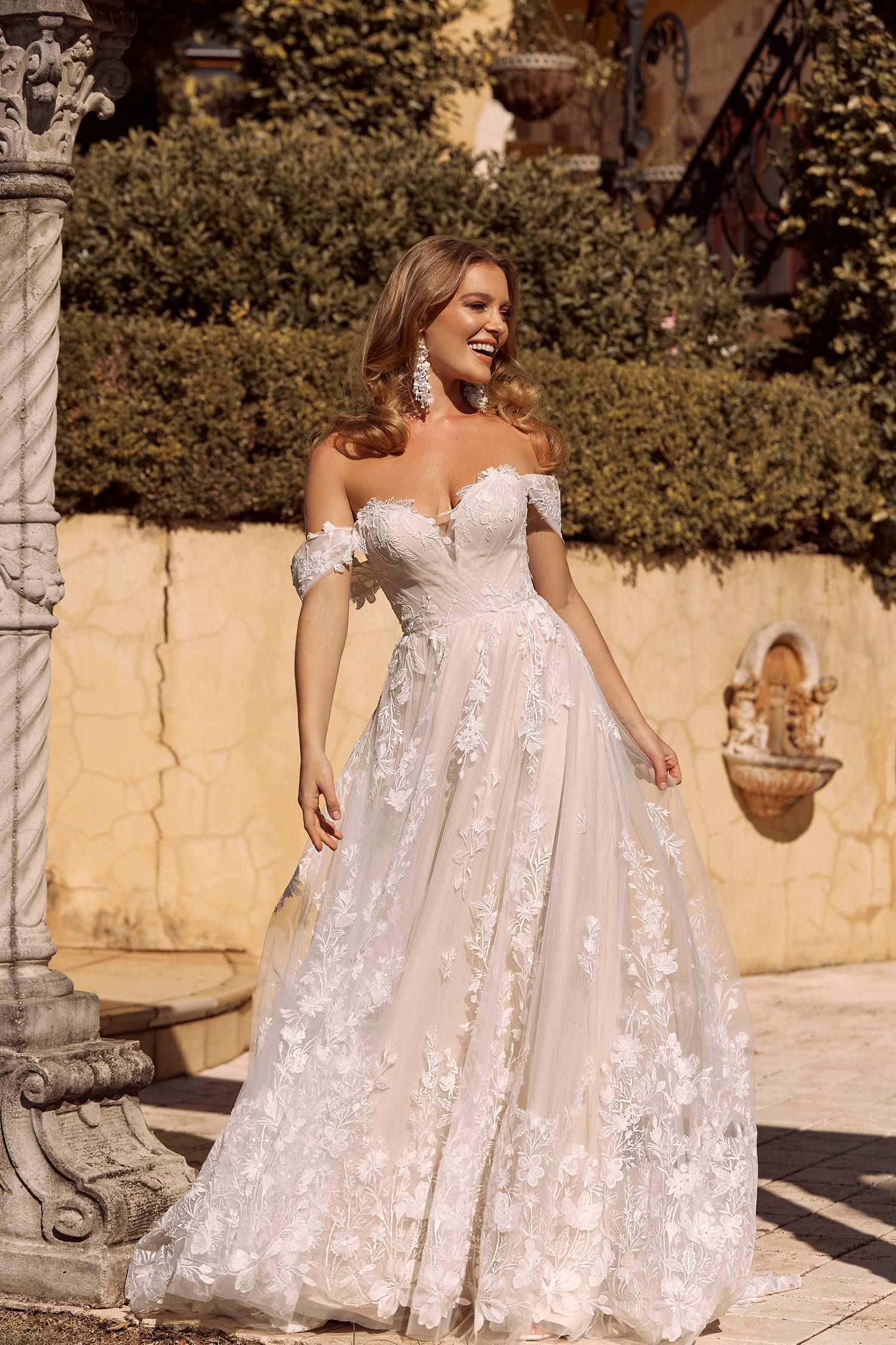 The image depicts a woman wearing an elegant off-the-shoulder wedding dress. The gown features a sweetheart neckline and intricate lace detailing throughout the bodice and skirt. The dress flows gracefully to the floor, creating a classic and romantic bridal look.