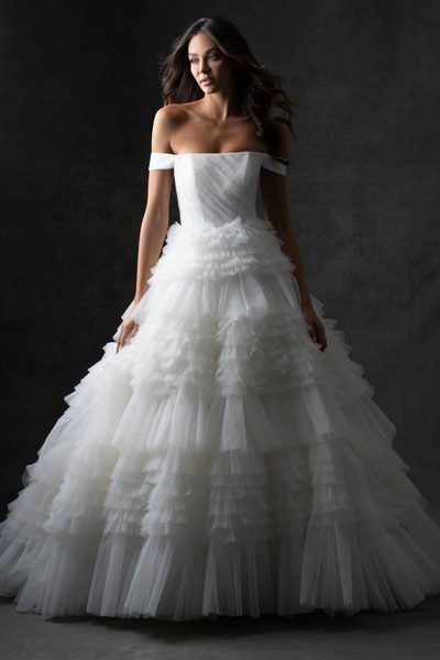 The image features a woman wearing a stunning, off-the-shoulder white wedding dress. The dress has a fitted bodice and a voluminous, multi-layered tulle skirt that creates a dramatic and elegant silhouette. The layers of tulle add texture and depth to the skirt, giving it a fairy-tale-like appearance. The woman is standing against a dark, neutral background, which makes the white dress stand out prominently. The lighting highlights the intricate details of the dress, emphasizing its luxurious and sophisticated design. The overall effect is one of timeless elegance and bridal beauty.