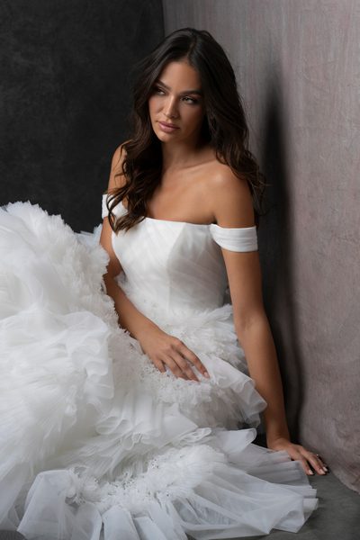 The image features a woman wearing a stunning, off-the-shoulder white wedding dress. Showing the bride sitting down in the layers of tulle. The dress has a fitted bodice and a voluminous, multi-layered tulle skirt that creates a dramatic and elegant silhouette. The layers of tulle add texture and depth to the skirt, giving it a fairy-tale-like appearance. The woman is standing against a dark, neutral background, which makes the white dress stand out prominently. The lighting highlights the intricate details of the dress, emphasizing its luxurious and sophisticated design. The overall effect is one of timeless elegance and bridal beauty.