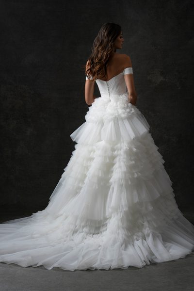 The image features a woman wearing a stunning, off-the-shoulder white wedding dress. Showing the back view of the gown. The dress has a fitted bodice and a voluminous, multi-layered tulle skirt that creates a dramatic and elegant silhouette. The layers of tulle add texture and depth to the skirt, giving it a fairy-tale-like appearance. The woman is standing against a dark, neutral background, which makes the white dress stand out prominently. The lighting highlights the intricate details of the dress, emphasizing its luxurious and sophisticated design. The overall effect is one of timeless elegance and bridal beauty.