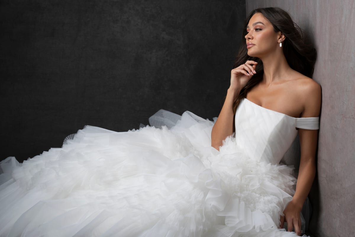 The image features a woman wearing a stunning, off-the-shoulder white wedding dress. The dress has a fitted bodice and a voluminous, multi-layered tulle skirt that creates a dramatic and elegant silhouette. The layers of tulle add texture and depth to the skirt, giving it a fairy-tale-like appearance. The woman is standing against a dark, neutral background, which makes the white dress stand out prominently. The lighting highlights the intricate details of the dress, emphasizing its luxurious and sophisticated design. The overall effect is one of timeless elegance and bridal beauty.