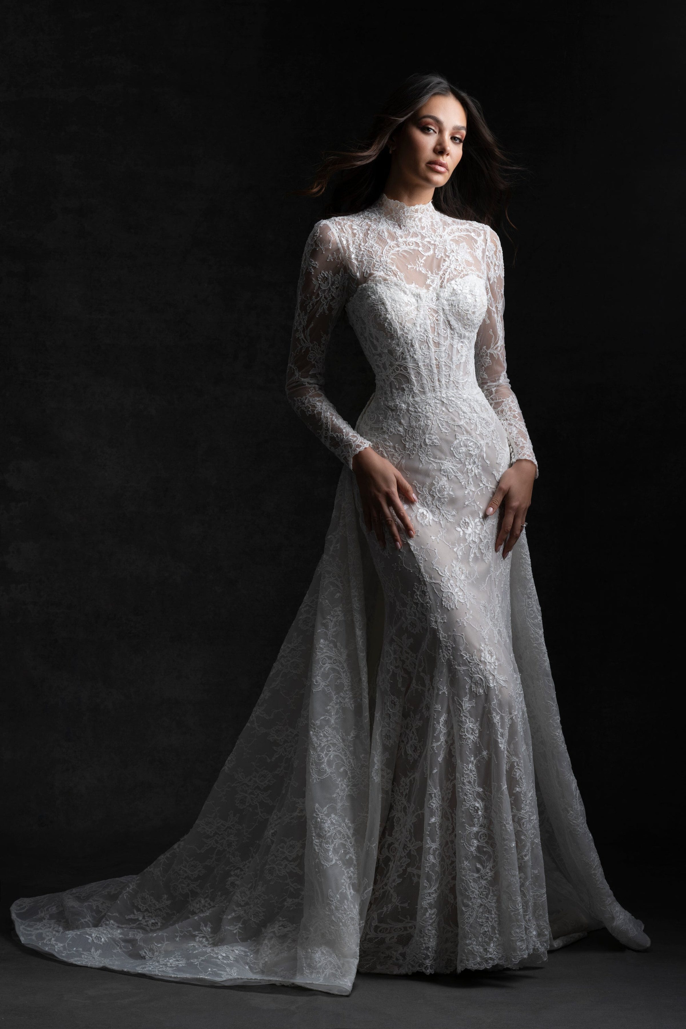 Bride in a high-neck, long-sleeved lace gown with a fitted silhouette and a long, elegant train, standing poised against a dark, moody backdrop.