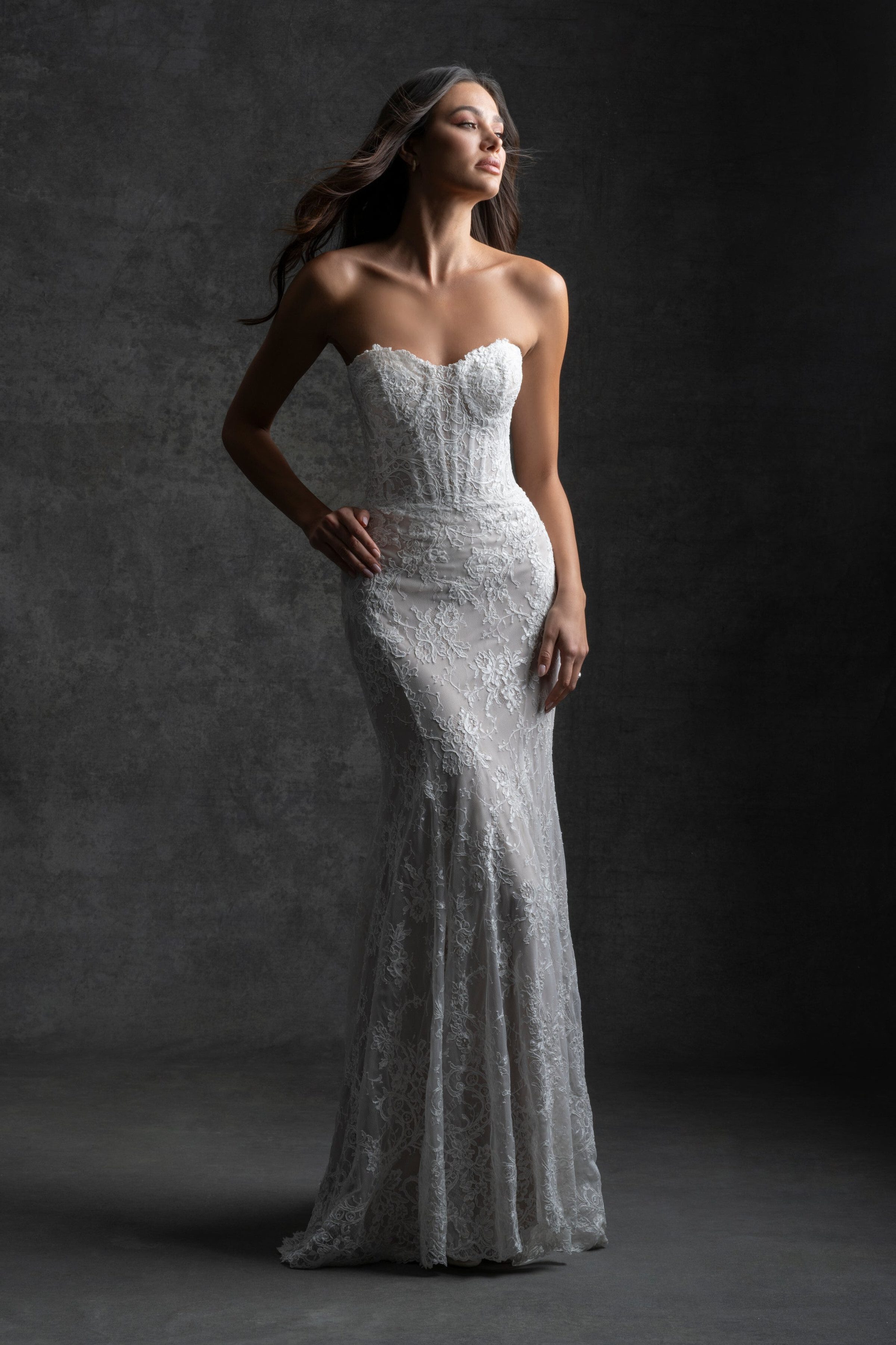 Bride in a strapless lace gown standing confidently with a hand on her hip, against a dark textured background.
