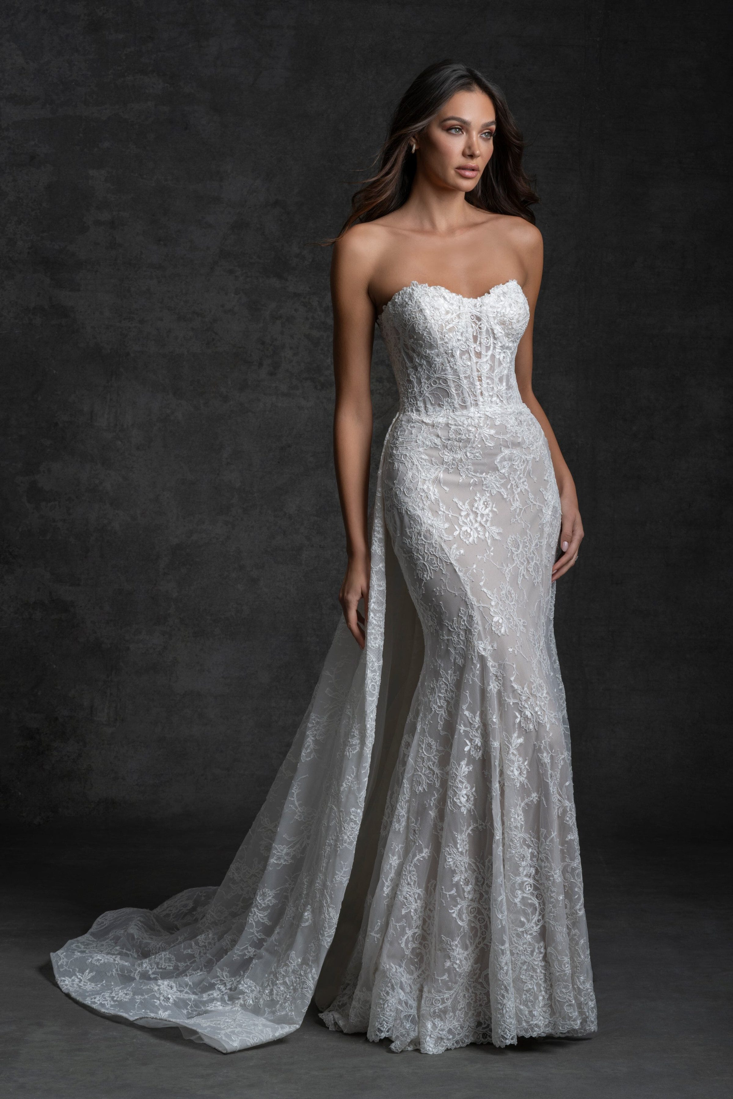 Bride in a strapless lace gown with a long train, standing elegantly against a dark textured background.