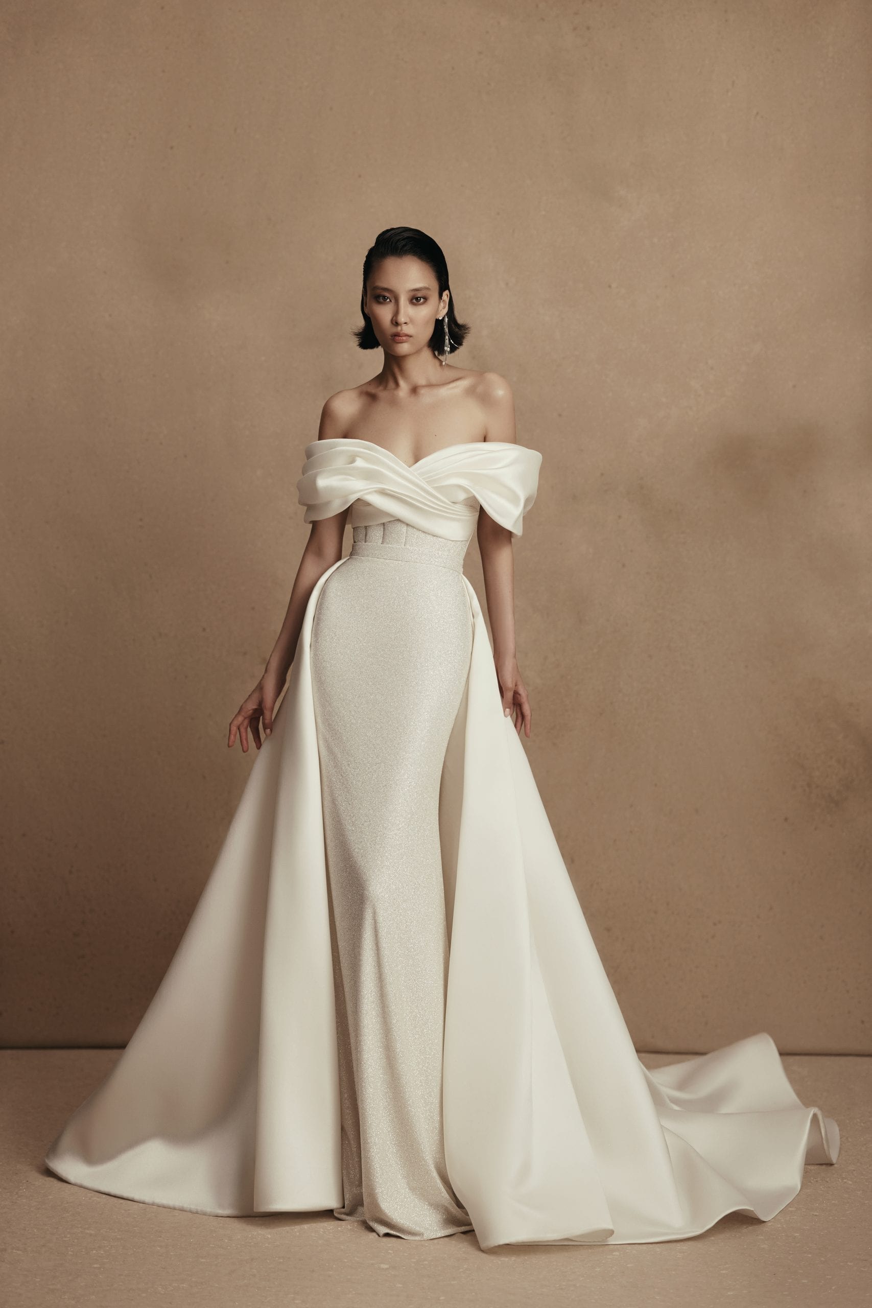 The image shows a bride wearing an elegant, off-the-shoulder white wedding dress. The gown features a draped design across the bodice, creating a flattering neckline that highlights the shoulders and collarbones. The bodice is fitted, cinching at the waist, and the dress flows into a full, voluminous skirt that extends into a long train, adding a dramatic and classic touch. The bride's hair is styled in an elegant updo, complementing the sophisticated and regal look of the gown. Her pose is poised and graceful, enhancing the overall elegance and timeless beauty of her appearance.