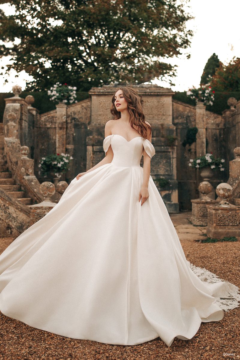 In a serene, historic garden, a bride embodies the grace and elegance of Disney's Belle. She is surrounded by ornate stone structures and lush greenery, creating a fairy-tale atmosphere. The bride's off-the-shoulder gown features delicate puff sleeves and a sweetheart neckline, exuding timeless romance. The voluminous skirt flows gracefully, adding a touch of magic to the scene. Her long, wavy hair and natural makeup highlight her ethereal beauty, making her look like a true princess from a classic tale.