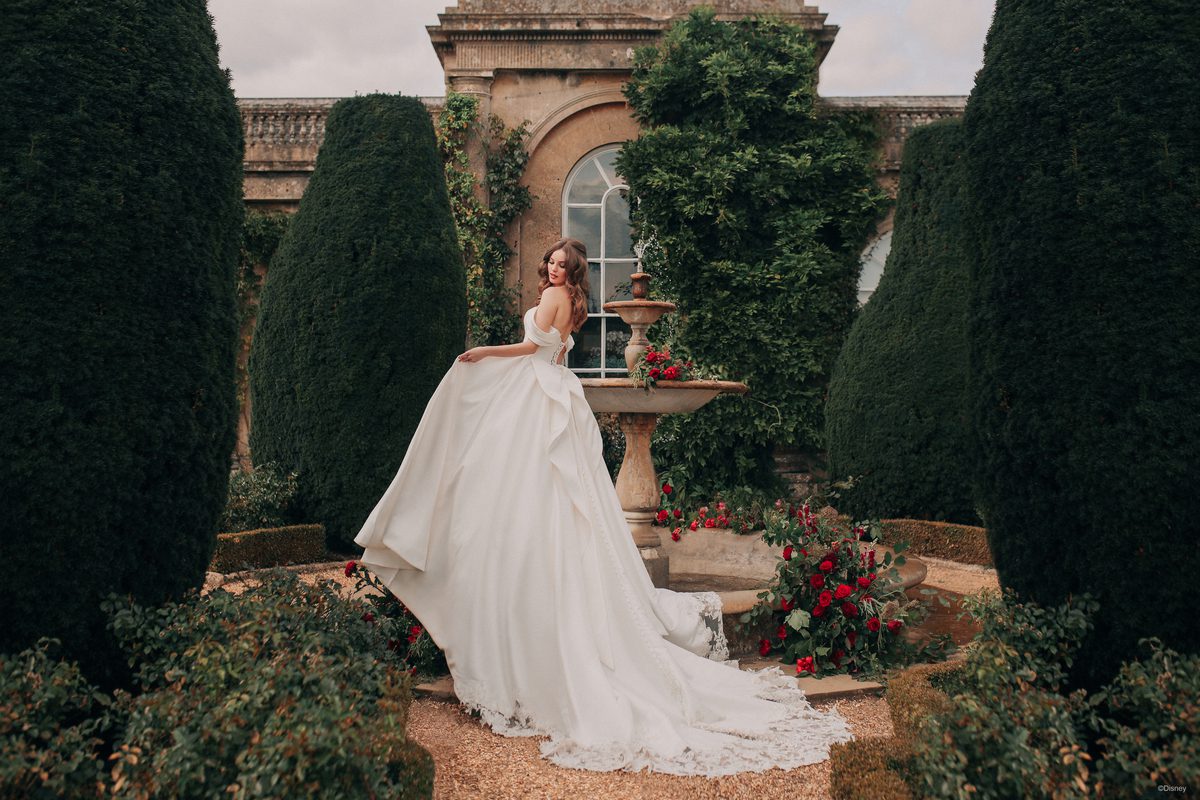 In a picturesque garden setting, a bride channels the elegance of Disney's Belle. She stands gracefully in front of a fountain, surrounded by manicured greenery and vibrant red roses. Her dress, reminiscent of Belle's iconic ball gown, features a voluminous skirt that cascades into a long, lace-trimmed train. The bodice is structured with a sweetheart neckline and delicate draping, adding a regal touch. Her soft, wavy hair and classic red lip complete the enchanting look, making her the epitome of fairy-tale romance.