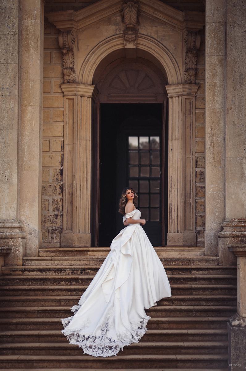 A bride stands regally on a grand staircase in front of an ornate, historical building, embodying the elegance of Disney's Belle. Her gown features off-the-shoulder puff sleeves, a fitted bodice, and a voluminous skirt that flows into a dramatic train adorned with intricate lace detailing. The soft waves of her hair frame her face, enhancing her serene and graceful expression. The architectural grandeur of the arched doorway and columns behind her add to the majestic, fairy-tale atmosphere, creating a scene that captures the timeless beauty and romance of a classic princess in a royal setting.