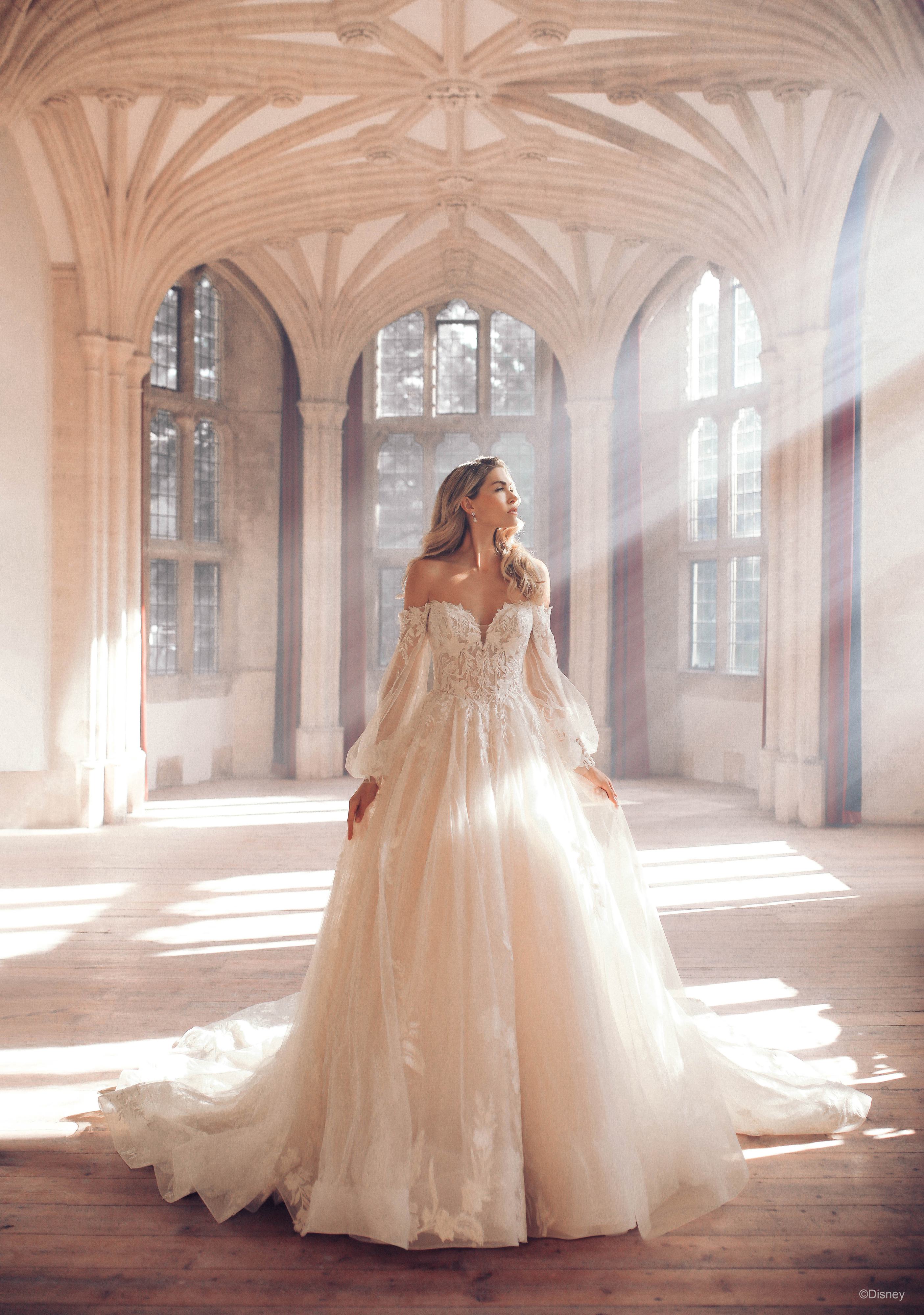 The image shows a woman wearing a stunning white wedding gown with a romantic and ethereal design. The dress features an off-the-shoulder neckline and long, sheer lace sleeves, adding a touch of elegance and sophistication. The bodice is fitted and adorned with intricate lace detailing, which extends slightly onto the full, voluminous skirt. The skirt is made of layers of light, airy fabric that flow gracefully to the floor and extend into a long, trailing train behind her. The overall look is dreamy and regal, perfect for a fairy-tale wedding. The woman's long hair is styled in loose waves, enhancing the romantic and timeless quality of the ensemble.