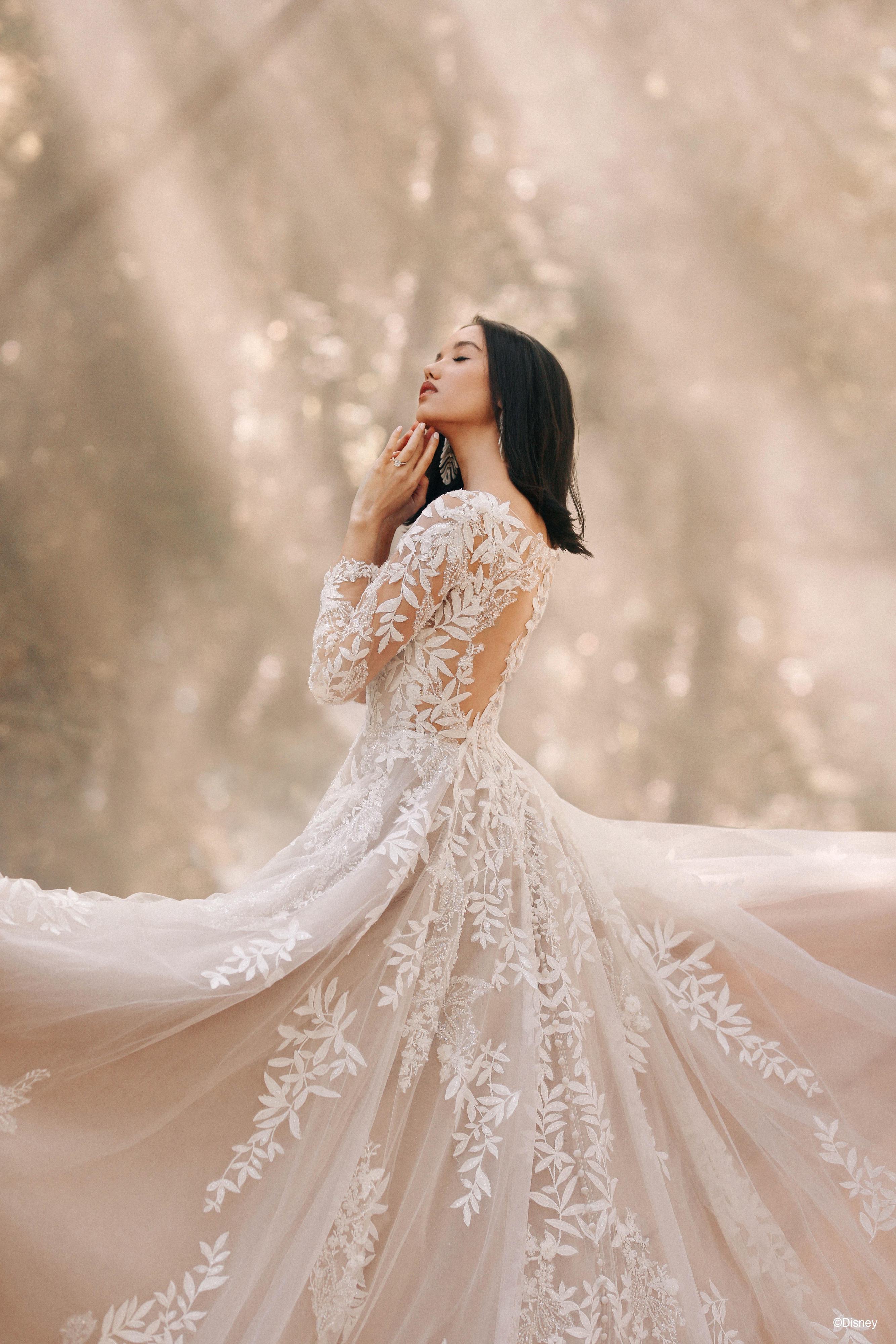 The image shows a woman wearing a delicate and elegant wedding gown with intricate lace detailing. The gown features long, sheer lace sleeves that add a touch of sophistication and romance. The bodice is fitted and also adorned with lace, complementing the sleeves. The woman's dark hair is styled straight and flows down her back. She appears to be in a contemplative or serene pose, with her eyes closed and her hands gently touching her face, enhancing the ethereal and timeless quality of the image. The overall effect is one of delicate beauty and elegance, perfect for a romantic wedding setting.