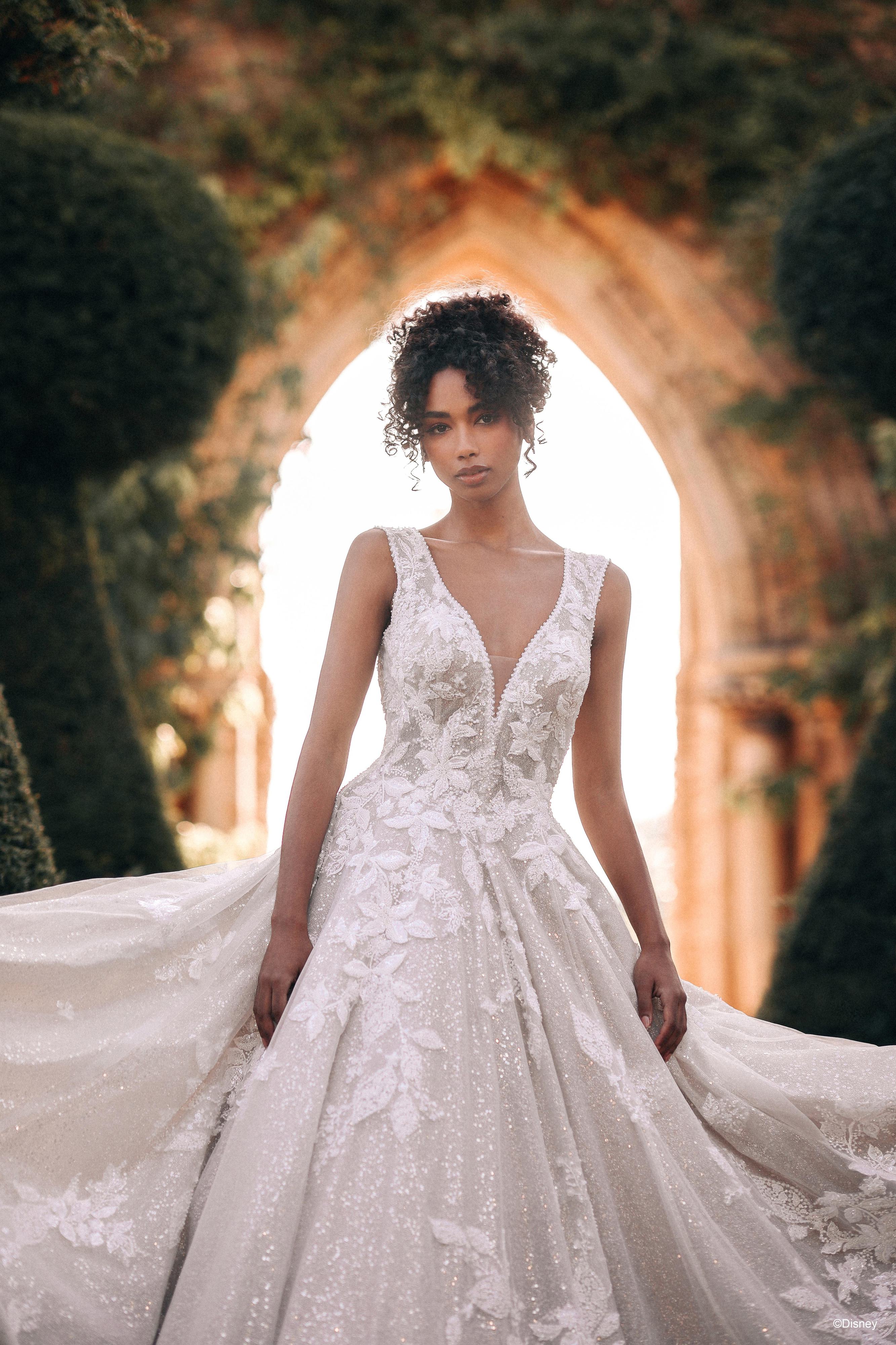 The image shows a woman wearing a stunning white wedding gown. The dress features a sleeveless design with a deep V-neckline, giving it a modern and elegant appearance. The bodice is intricately adorned with lace detailing, adding a touch of romance and sophistication to the gown. The skirt is full and voluminous, flowing gracefully from the fitted waist and creating a classic ballgown silhouette. The fabric of the skirt is rich and textured, with layers adding depth and grandeur. The woman's hair is styled in a chic updo, and she is wearing a delicate headpiece, enhancing the sophisticated and regal quality of the dress. The overall effect is one of timeless beauty and elegance, perfect for a fairy-tale wedding.