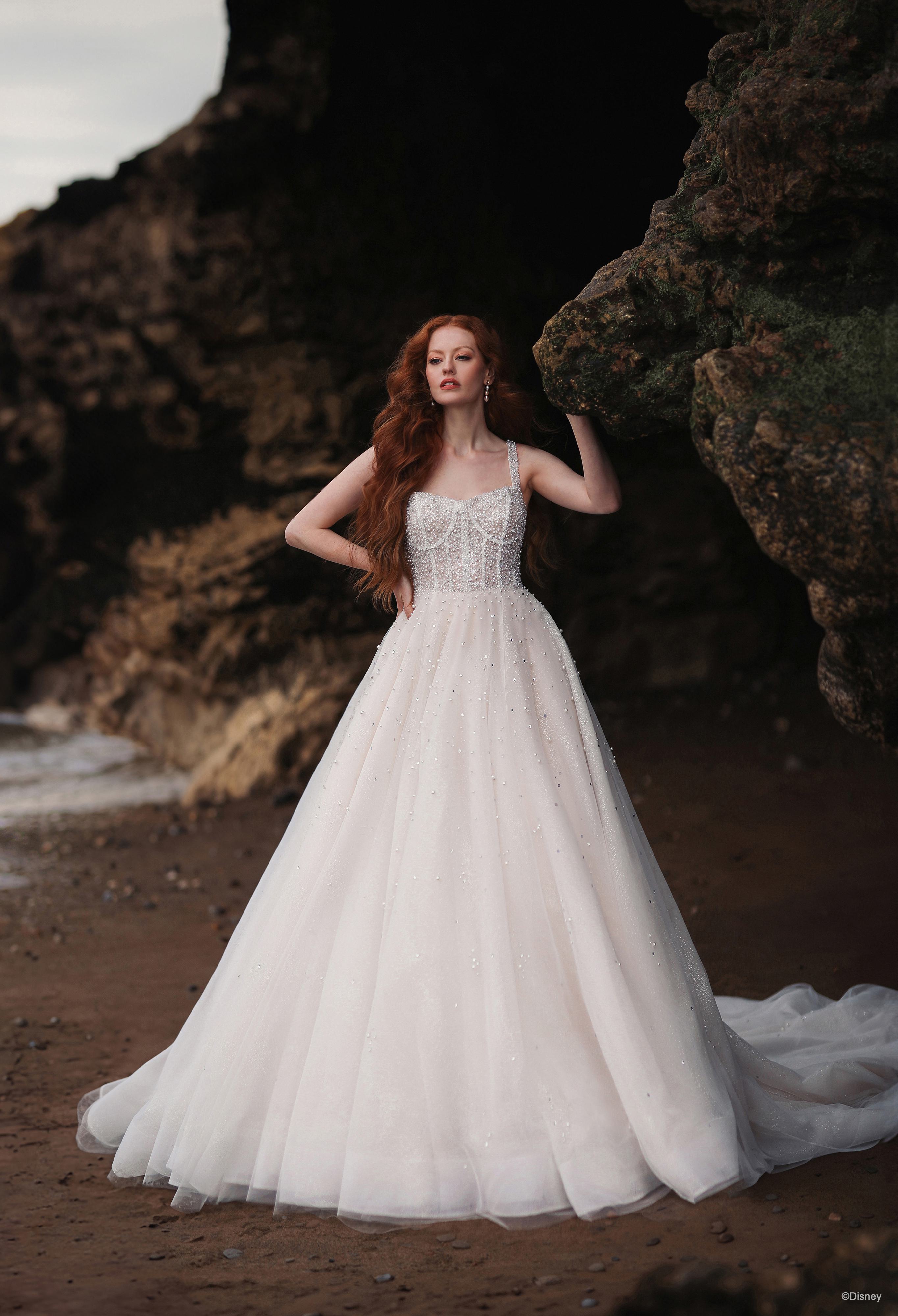 The image shows a woman wearing a stunning white wedding gown. The dress features a strapless, fitted bodice adorned with intricate lace and beading, creating a delicate and elegant appearance. The skirt is full and voluminous, made of layers of soft, airy fabric that flow gracefully to the floor, giving the dress a classic and romantic silhouette. The gown extends into a subtle train that adds to its elegance. The woman's long red hair is styled in loose, flowing waves, enhancing the romantic and ethereal quality of the gown. The overall look is timeless, sophisticated, and perfect for a fairy-tale wedding.