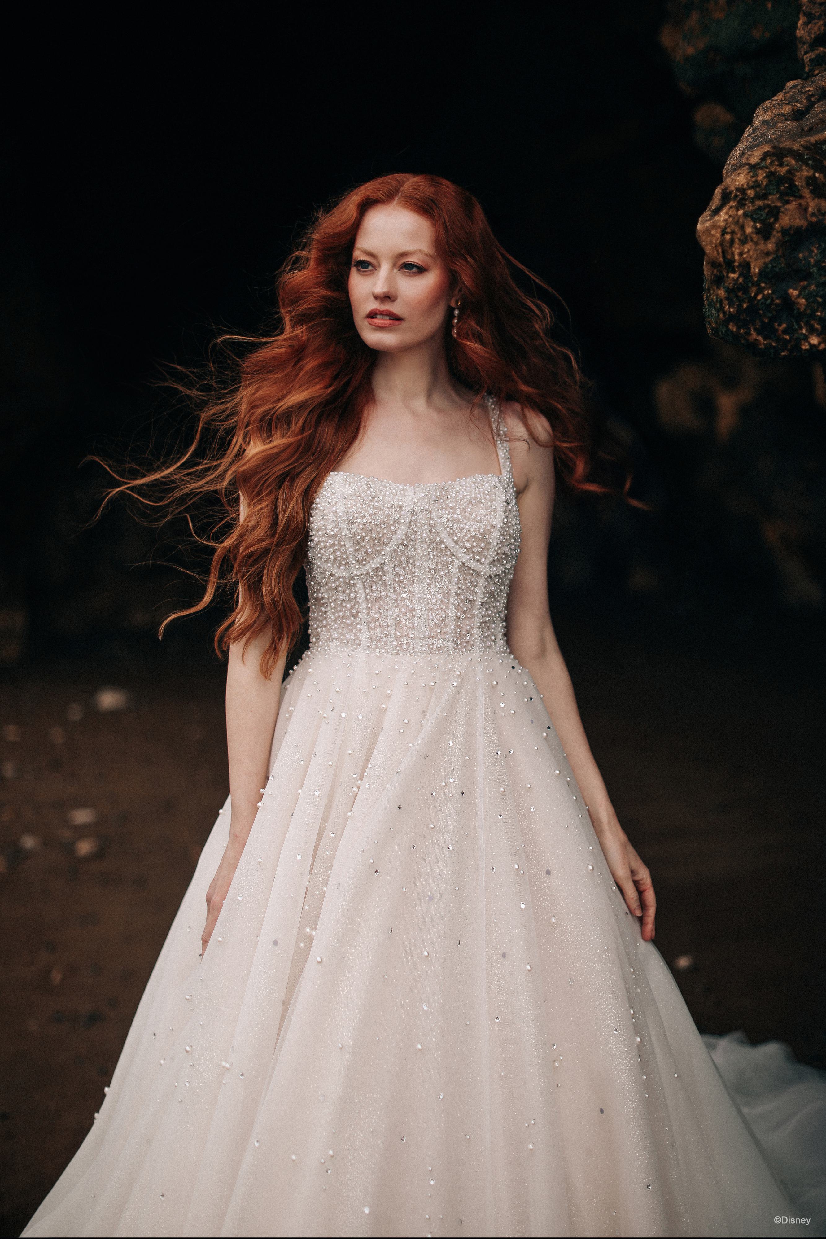 The image shows a woman wearing a beautiful and elegant white wedding gown. The dress features a strapless, fitted bodice adorned with intricate beading and lace detailing. The skirt is full and voluminous, made of layers of light, airy fabric that flow gracefully to the floor, creating a classic and romantic silhouette. The gown has a subtle sparkle, adding a touch of glamour to the overall look. The woman's long red hair is styled in loose, flowing waves, complementing the romantic and ethereal quality of the dress. The overall effect is one of timeless beauty and sophistication, perfect for a fairy-tale wedding.