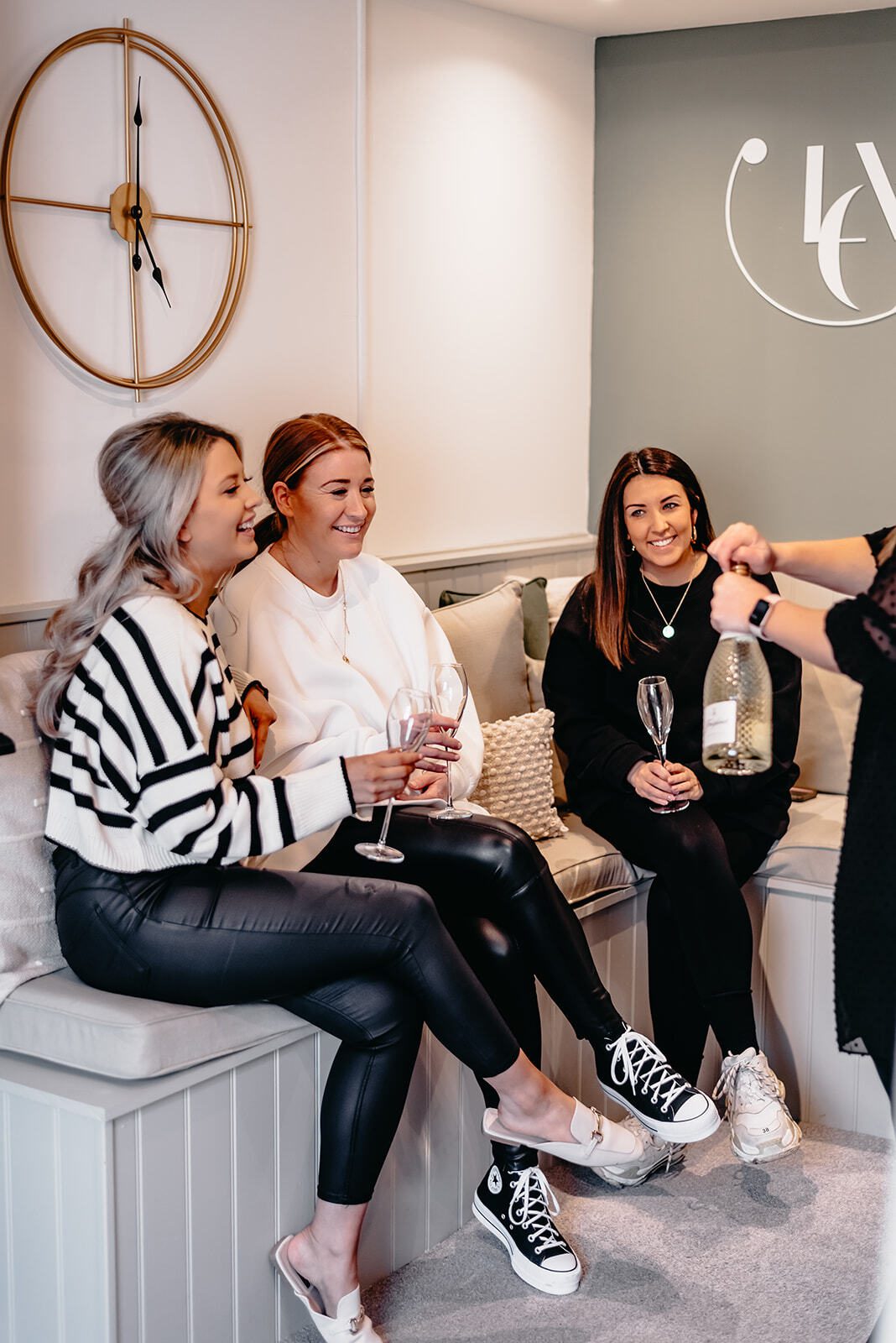 Three women sitting on a cushioned bench, smiling and holding wine glasses, while a fourth person pours a drink, in a cozy room with a modern clock on the wall.