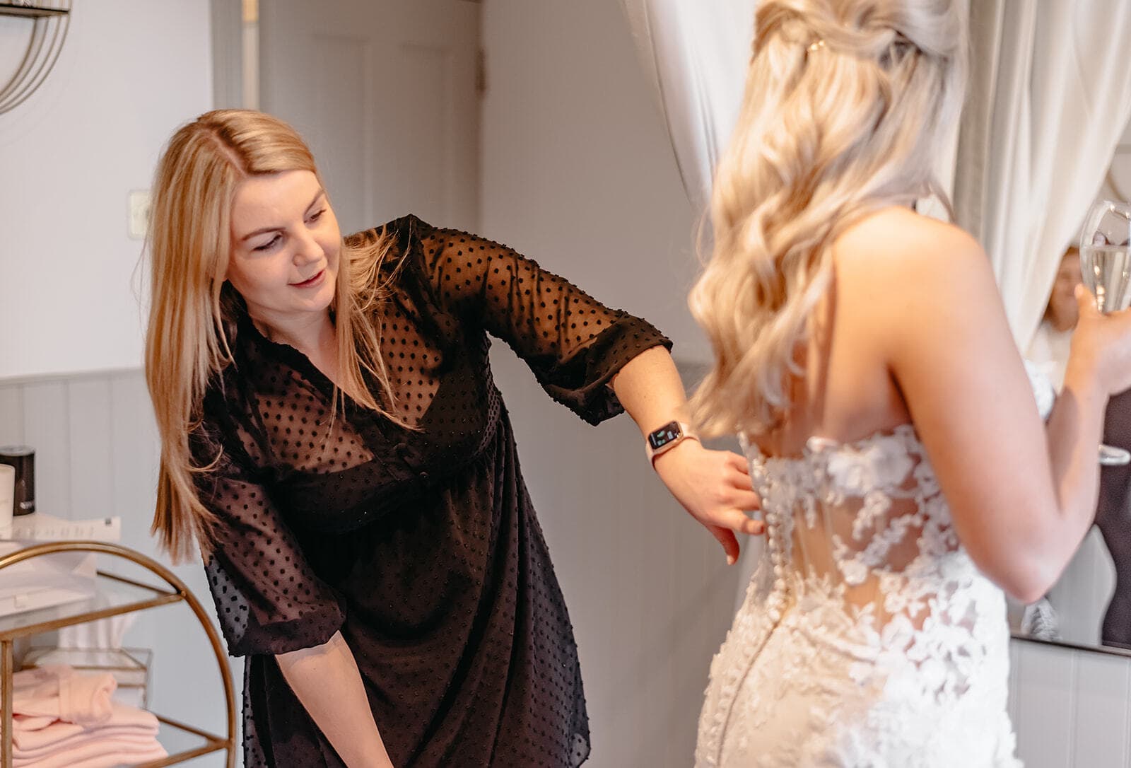 Woman in a black polka dot dress assisting a bride with her intricate lace gown, in a bright room with light curtains and a champagne glass in the bride's hand.
