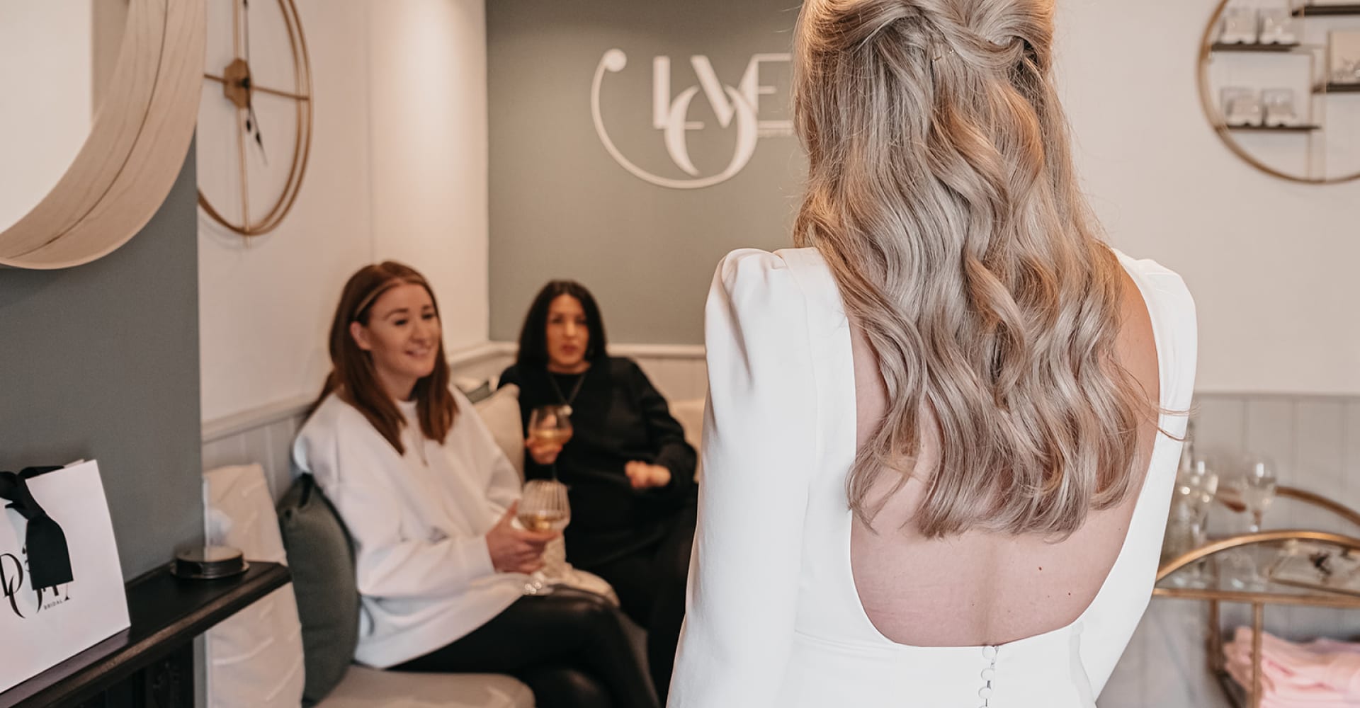 The image shows the interior of a bridal boutique where a bride-to-be is trying on a wedding dress. The focus is on the back of the bride, who has long, wavy blonde hair and is wearing a white dress with an open back. She is facing two women seated on a couch, who appear to be giving her feedback. The women are dressed casually, one in a light-colored sweater and the other in a dark top, and they are smiling and engaging in conversation with the bride. The boutique has a cozy and inviting atmosphere, with a mix of light and dark tones in the decor. On the wall behind the seated women, there is a decorative sign with the letters "YES" and some circular mirrors and shelves displaying various items.